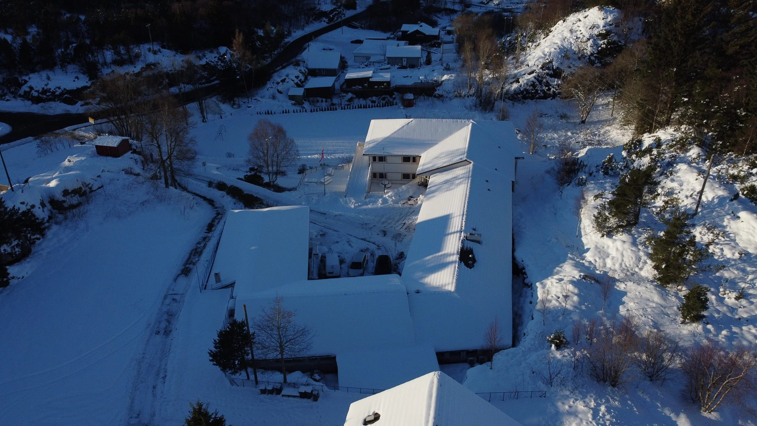 Fjernbilde av en hvit snødekt bygning med flere kjørebaner og trær rundt, omgitt av snø og vinterlandskap.