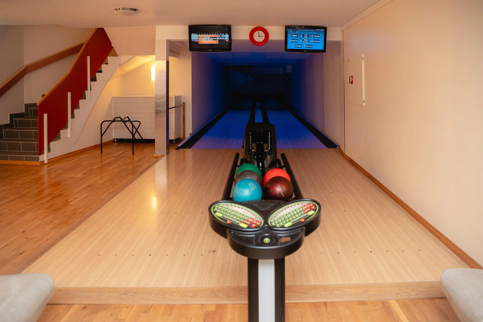 Innendørsscene av en bowlinghall med bowlingkuler og en klubb under, samt informasjonsskjermer og en klokke på veggen.