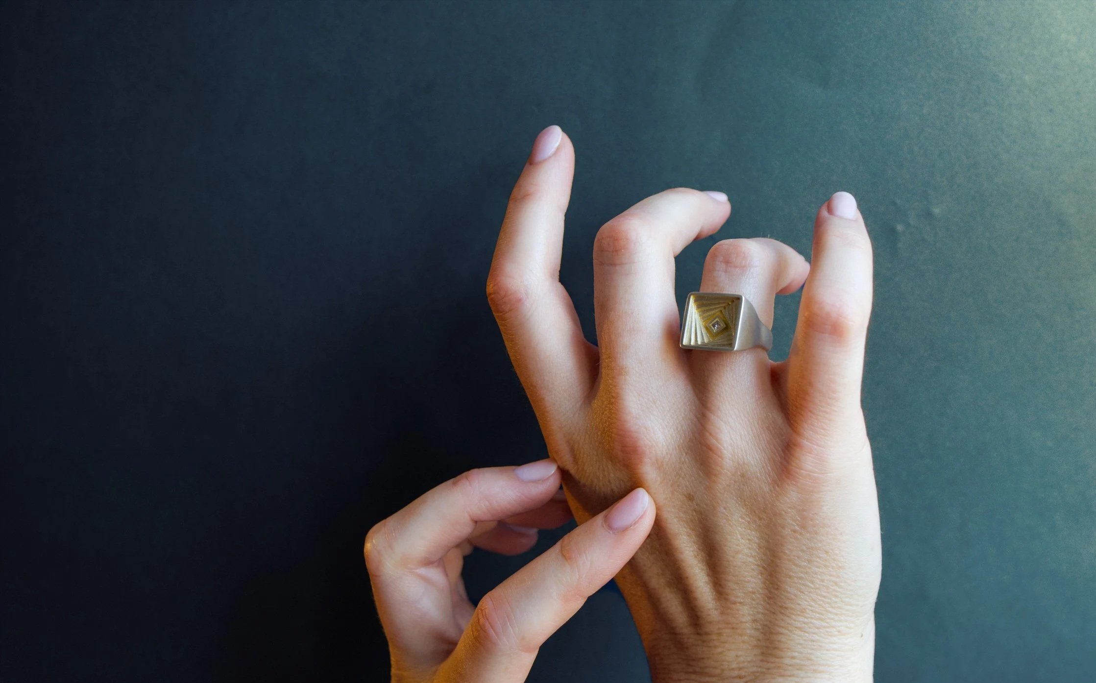 A hand wearing a large silver ring with a geometric design, holding another hand with fingernails slightly visible, against a dark background.