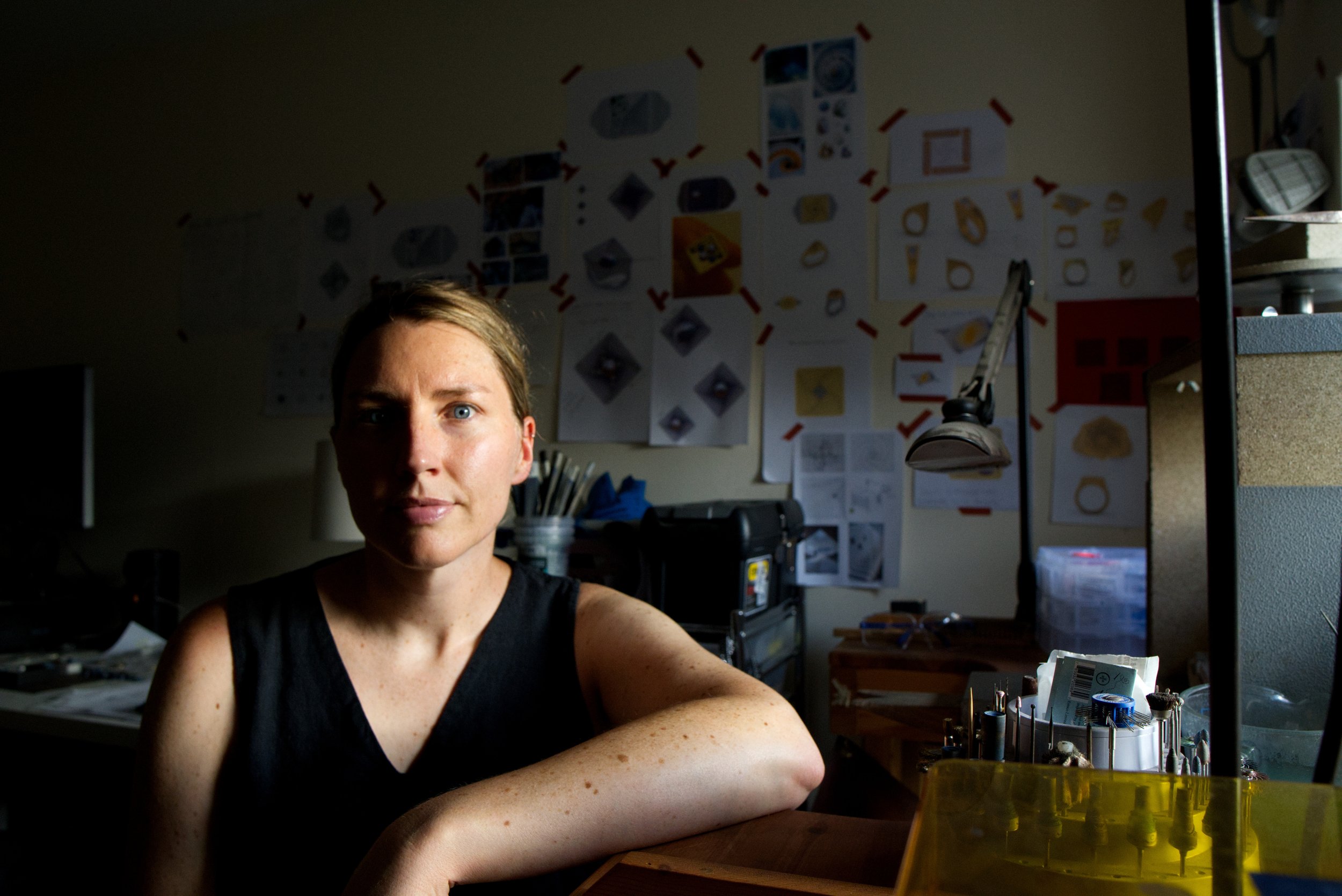 A woman with short brown hair and blue eyes sitting at a cluttered workbench in a workshop or laboratory, with various tools, parts, and papers on the wall behind her, and a computer monitor on her left.