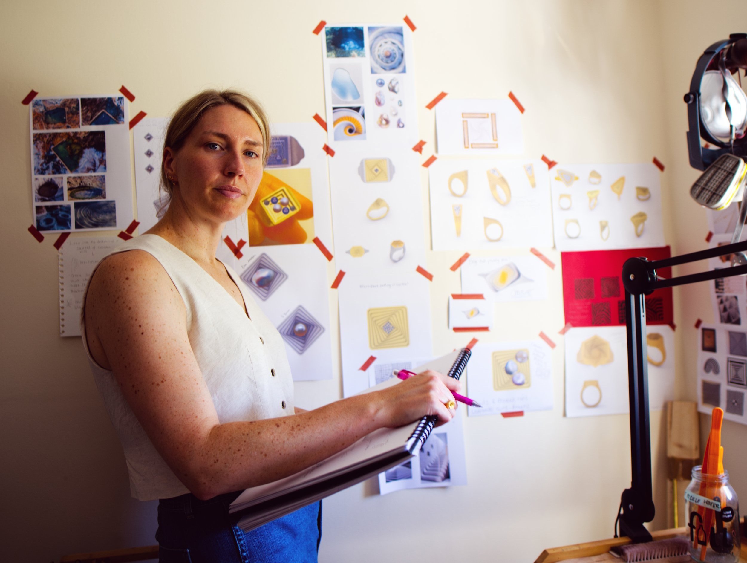 Beth Chesser stands in front of a wall covered with printed images and notes, holding a notebook and a pink pen, with a large studio light on her right.