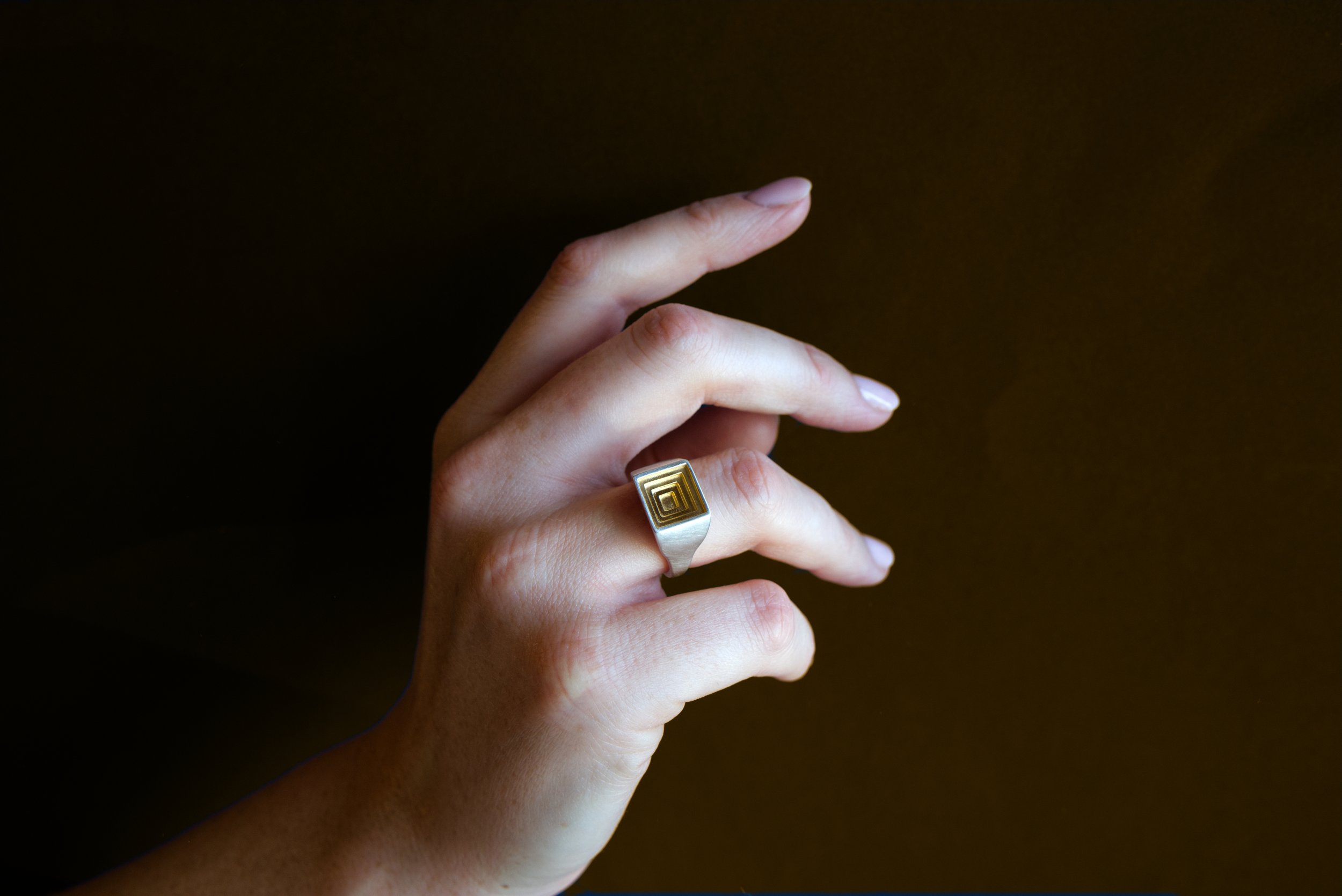 A person’s hand with a gold geometric patterned ring, showing fingers and part of the palm against a dark background.