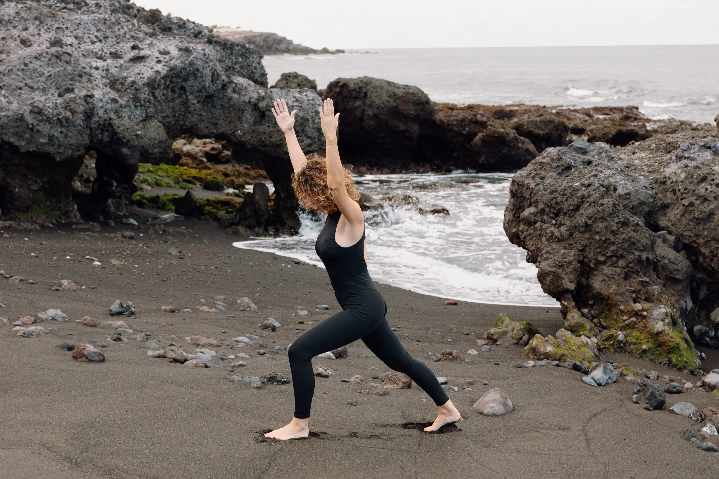 Persona practicando yoga en la playa frente a rocas, con el mar al fondo.