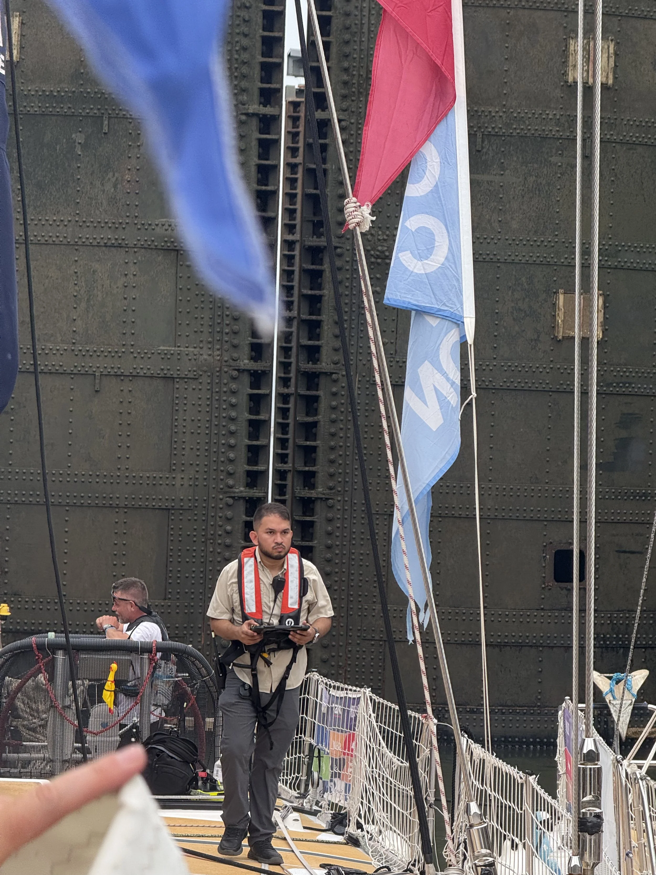Sailors, a pilot and crew members standing on a boat deck with flags and rigging, with a large dark metal wall in the background.