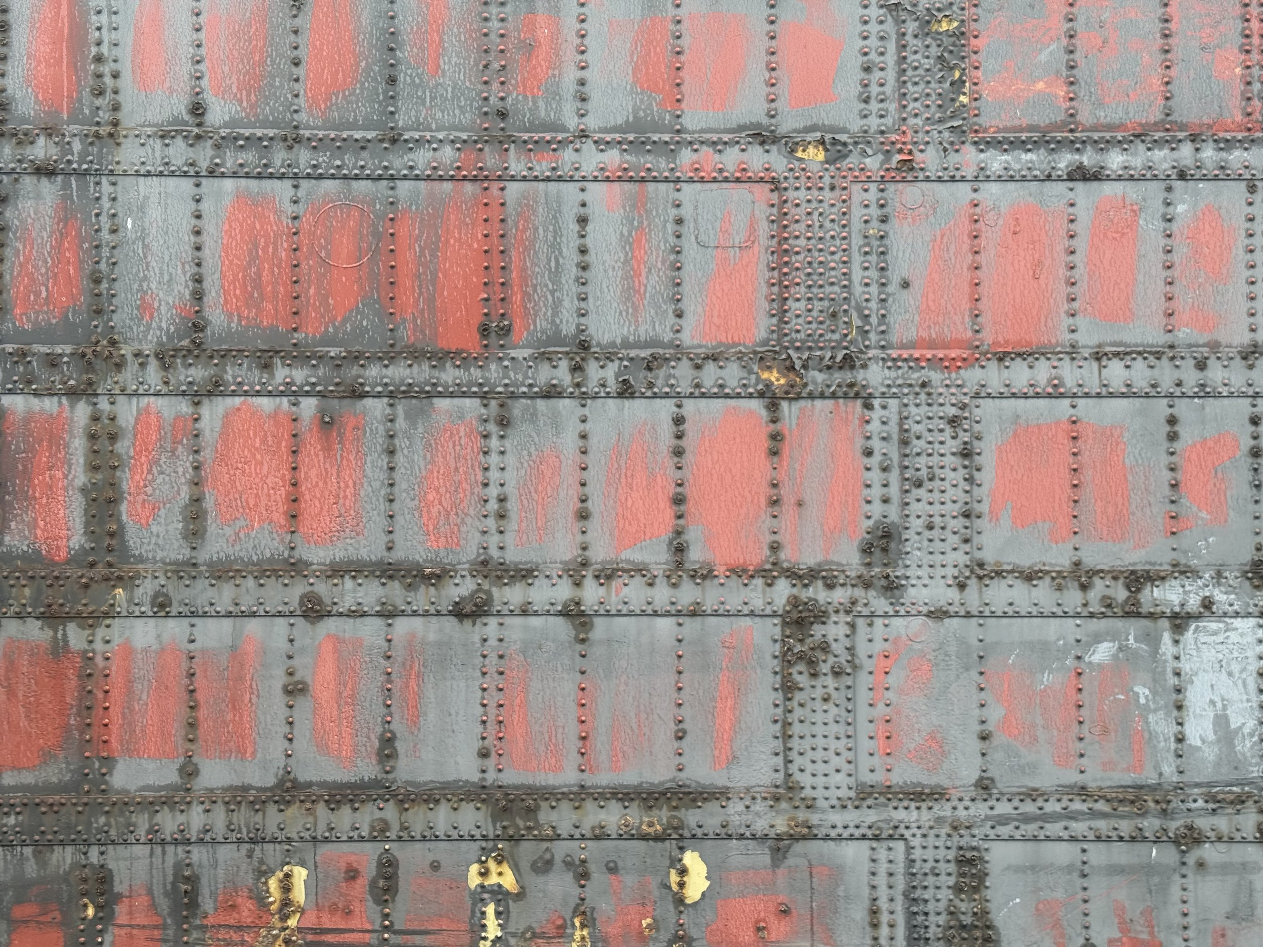 Close-up of an old metal surface of a lock door with rivets, painted in gray and red, showing peeling paint, rust, and signs of wear.