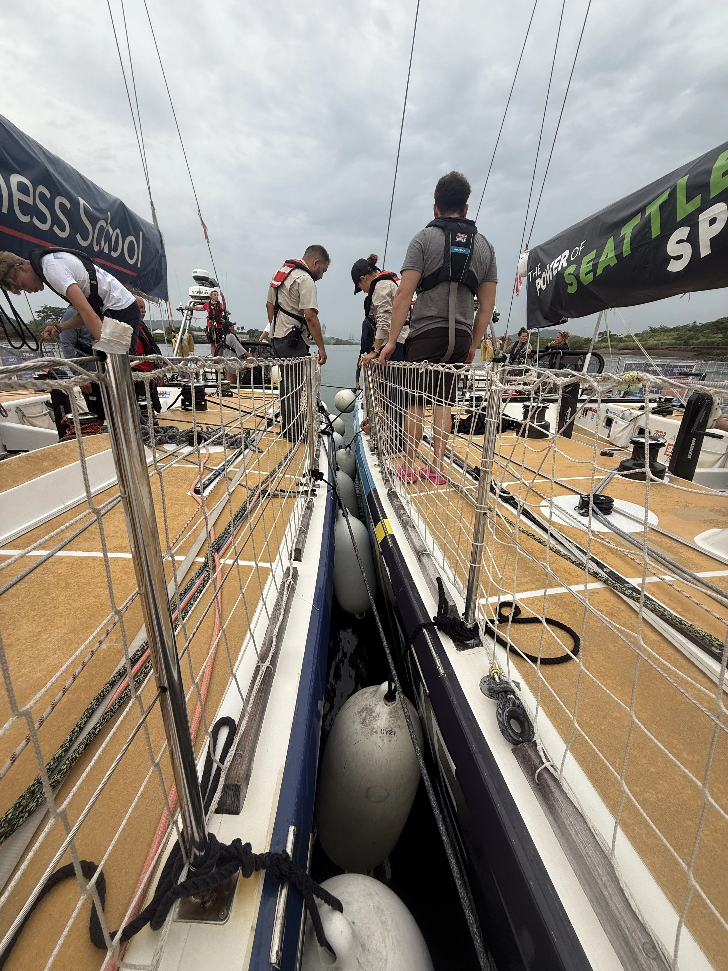 A group of people on a sailboat preparing for getting in the locks of the Panama Cnal, standing on the deck surrounded by safety rails and equipment, with water and trees visible in the background under a cloudy sky.