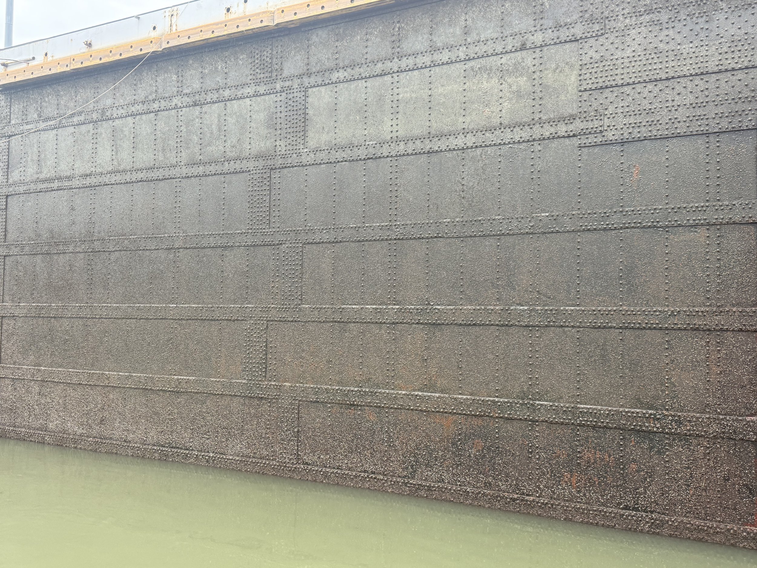 Close-up of a section of dark gray metal of one of the lock doors of the Panama Canal with rivets, part of a larger structure, with green painted surface below.