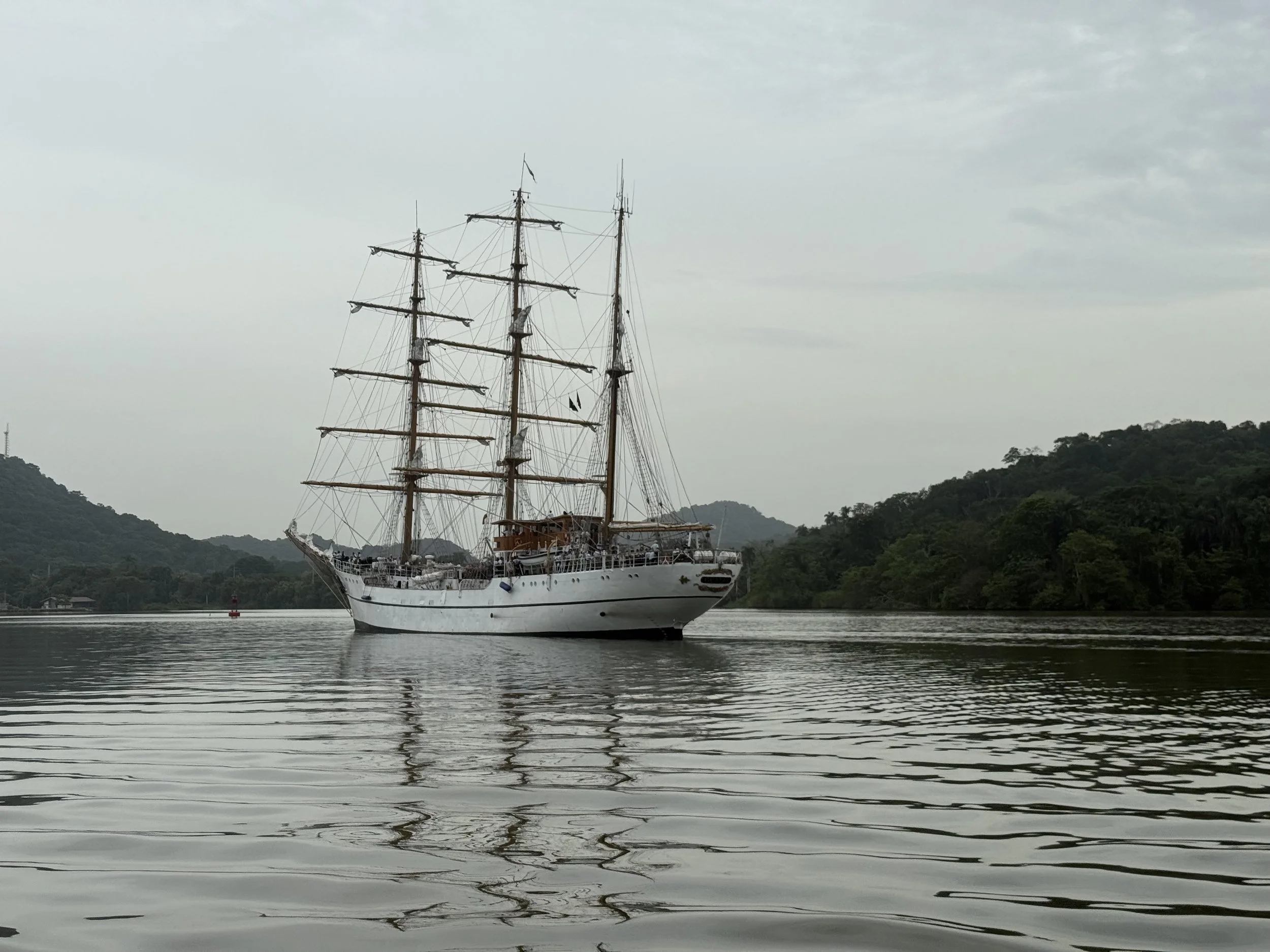 A large white sailing ship with multiple masts and rigging is anchored in calm waters near a lush, green hillside. Overcast sky in the background.