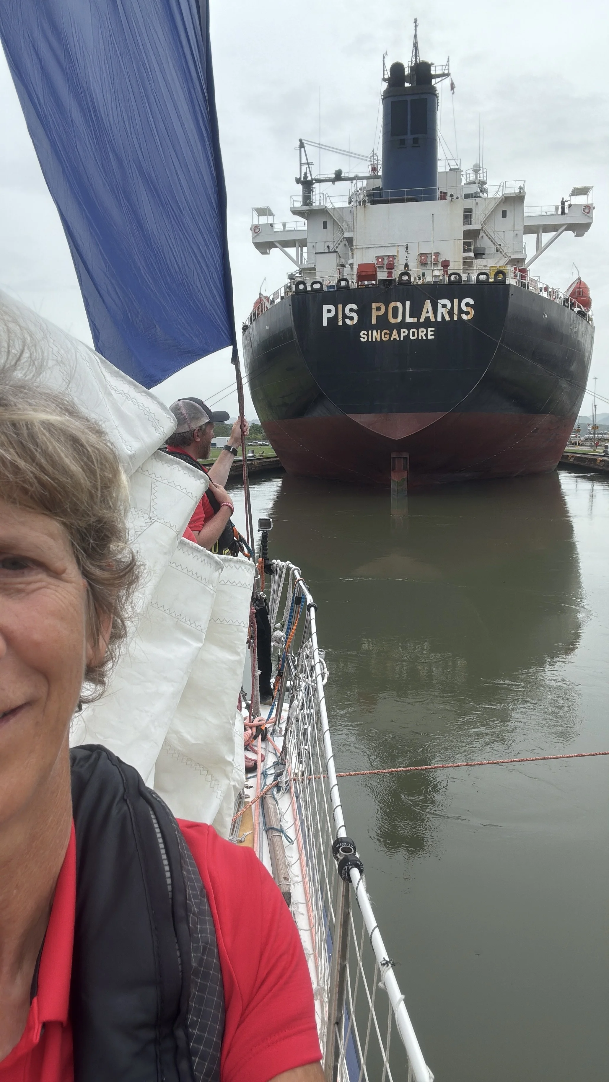 A woman on a boat holding a blue flag, with a large ship named PIS POLARIS from Singapore docked in the water in front of the boat.