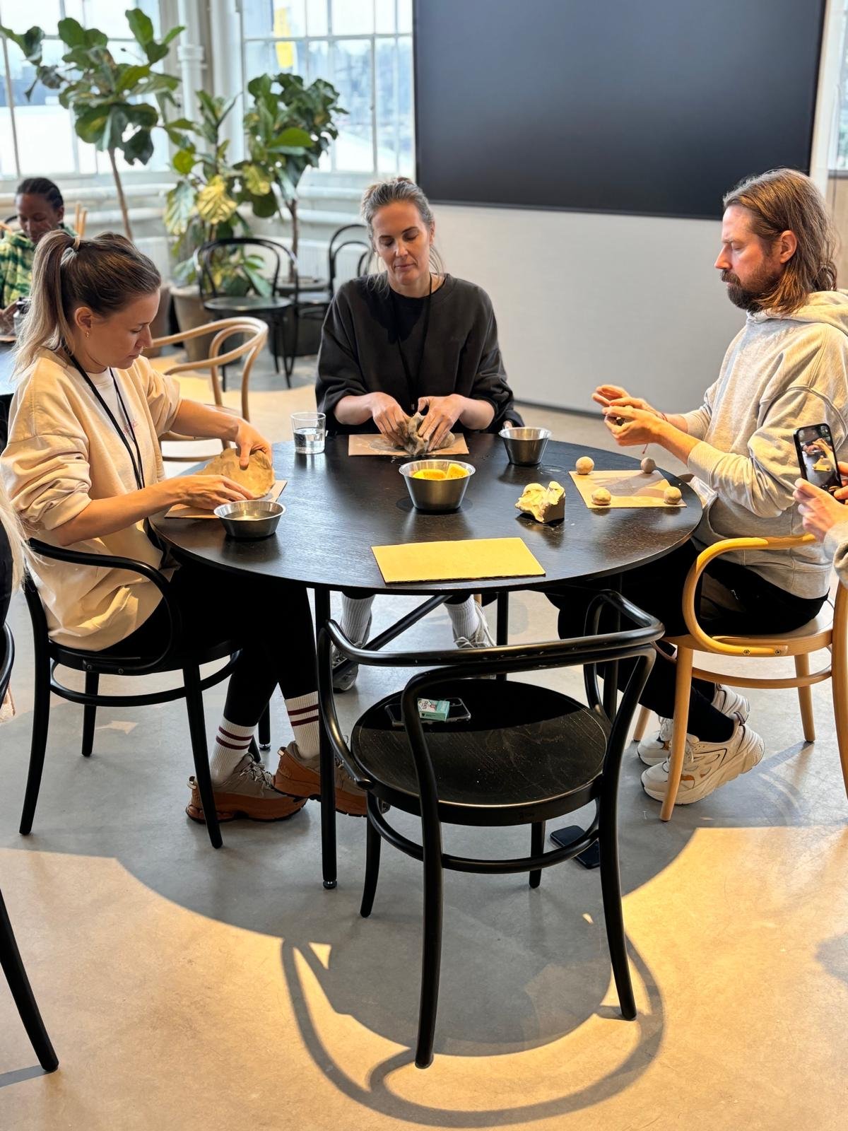 Tre personer som gör degknyten vid ett runt bord, med ingredienser och deg. Ett svart stort skärm i bakgrunden.