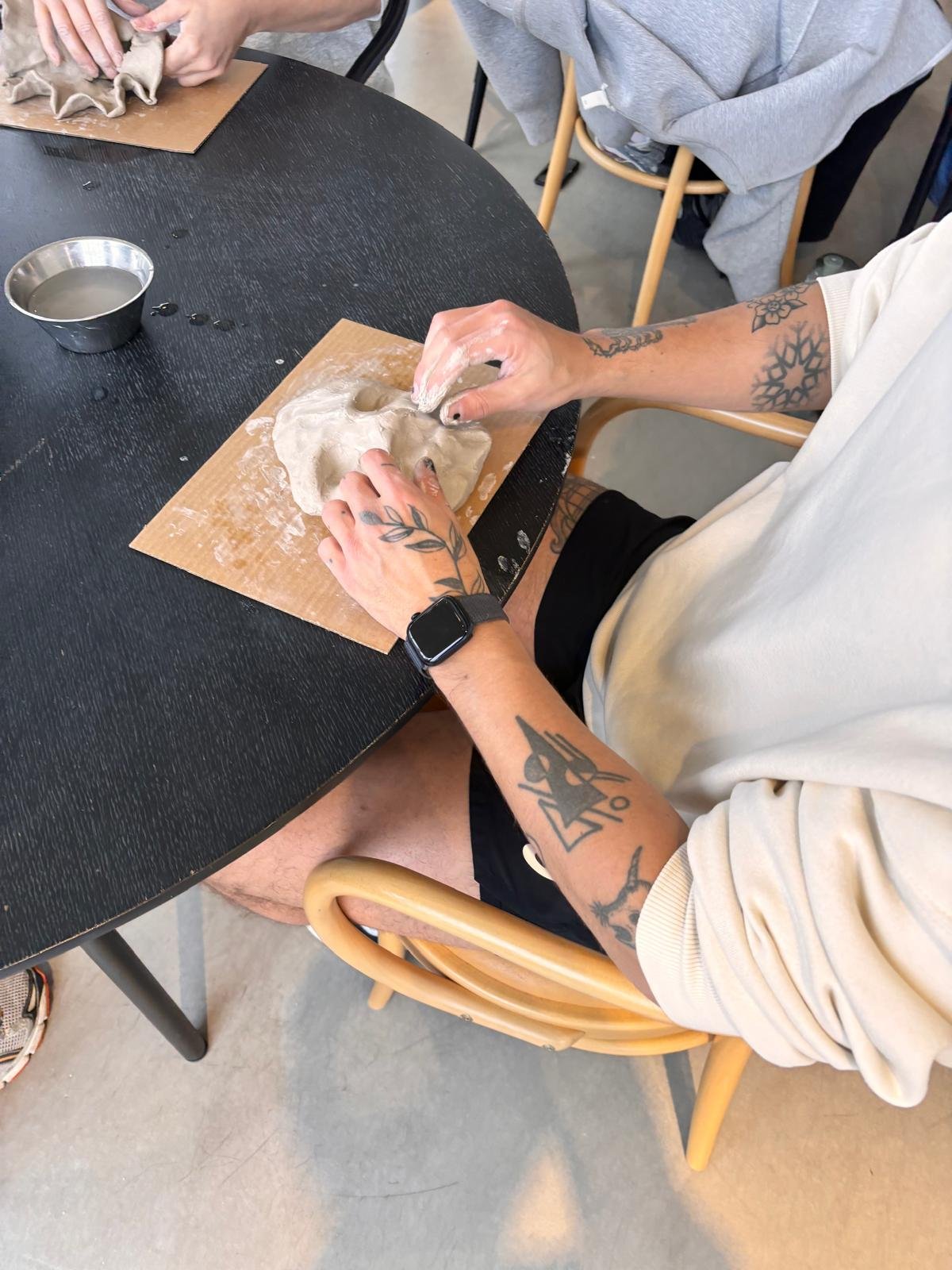 Person som rör på en leradfas med ett ansiktsmotiv, sittande vid ett svart bord, andra händer arbetar med samma lera i bakgrunden, på en trästol.