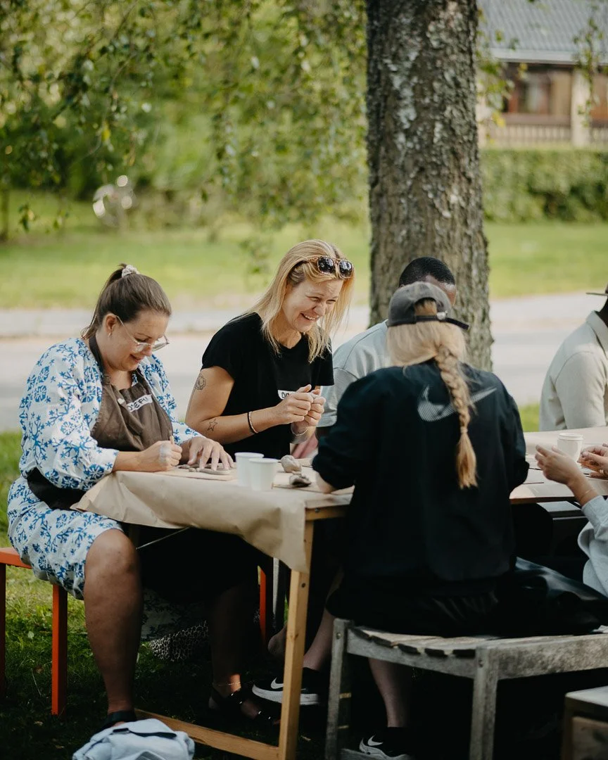 Grupp människor som skrattar och pratar vid ett utomhusbord under ett träd.