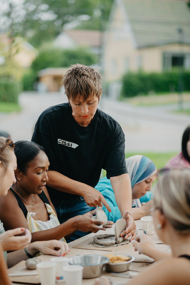 En grupp människor deltar i en keramikkurs utomhus, med en instruktör som visar hur man formar lera.