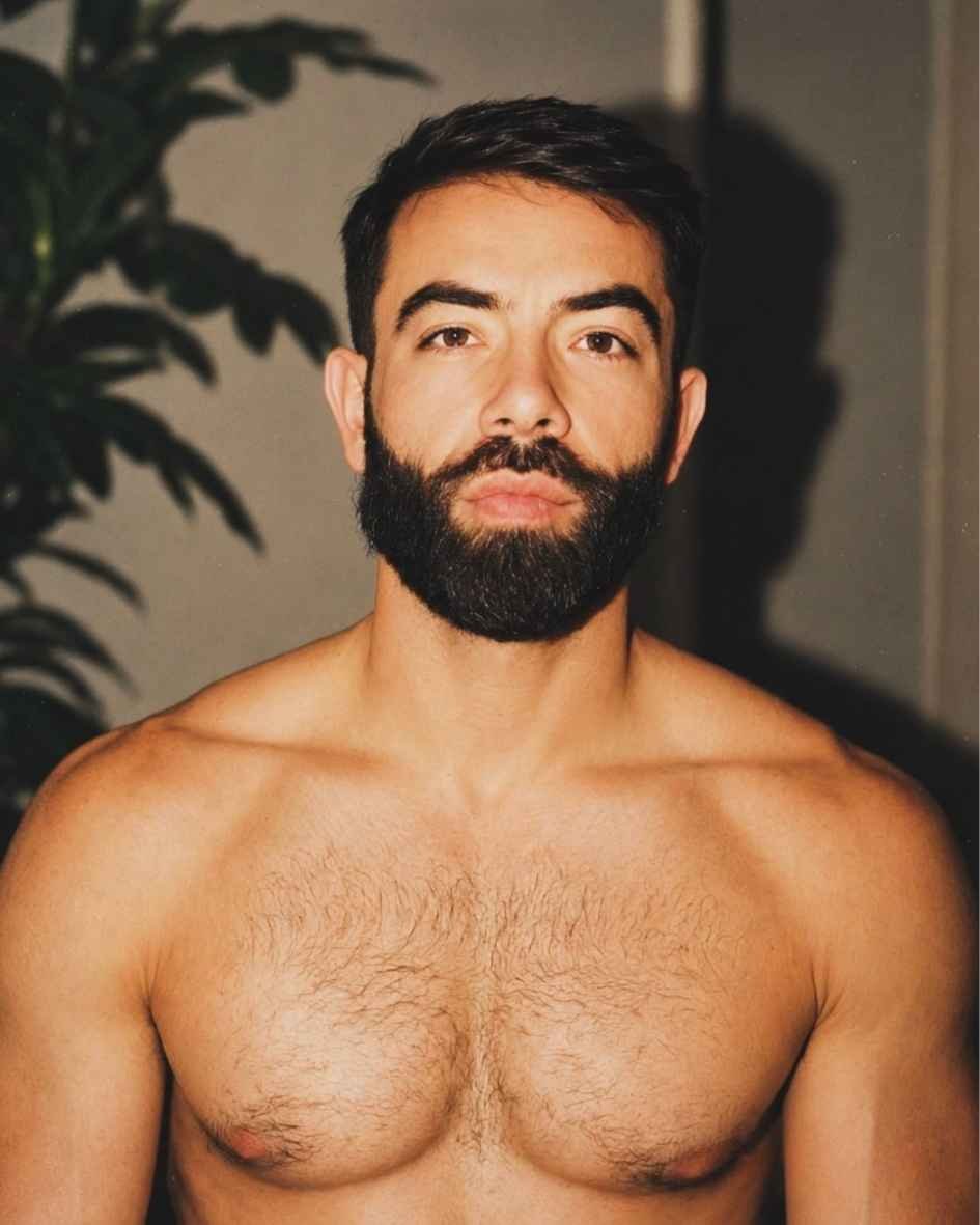A shirtless man with a beard and short dark hair looking directly at the camera, in front of a gray wall and some green plant leaves.
