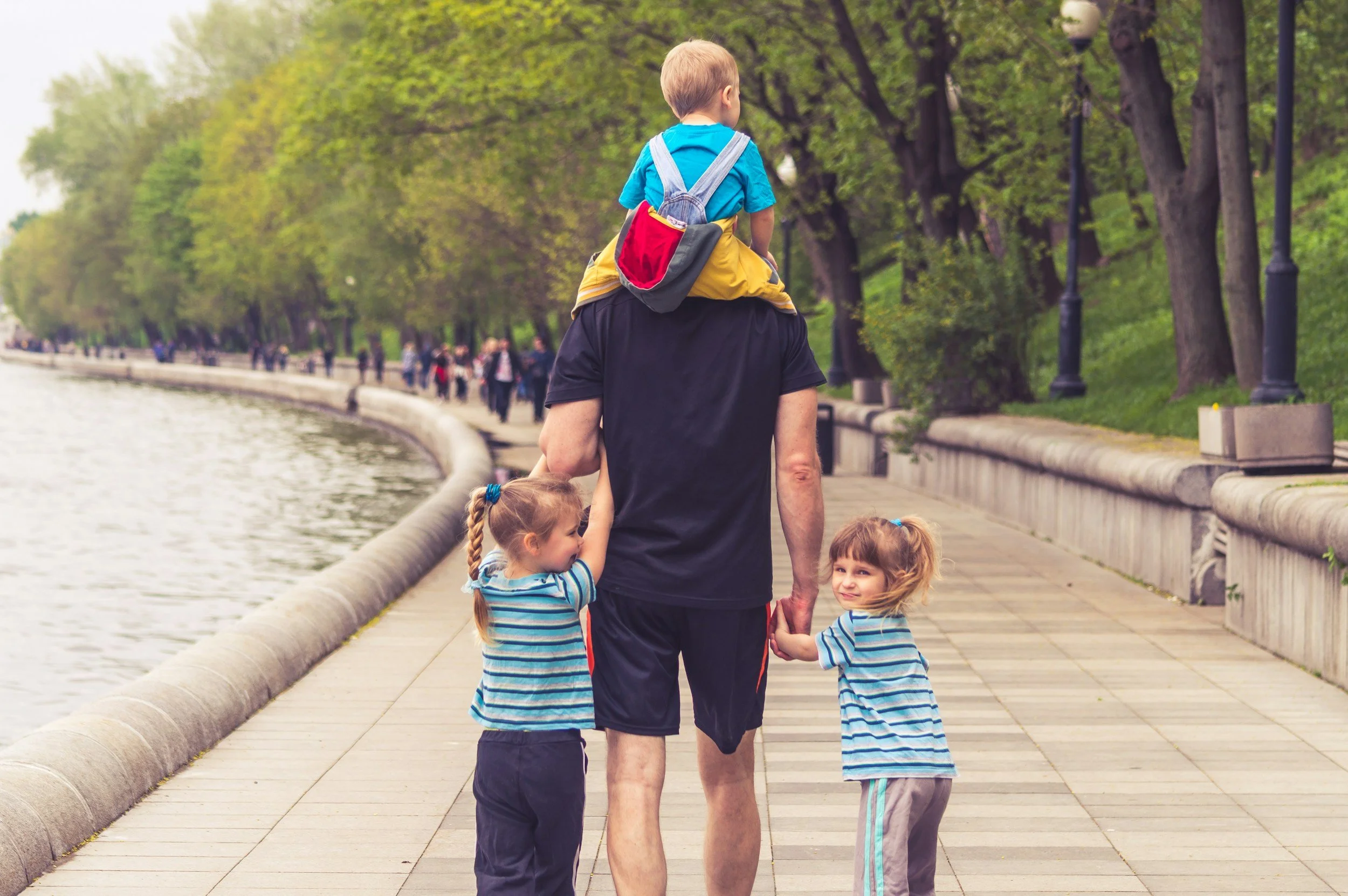 A man walking along a riverside walkway with two young girls, one sitting on his shoulders, in a park with green trees and people in the background.