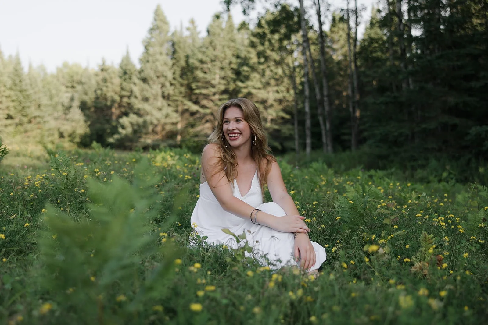 A young woman with long, wavy blonde hair sitting on a grassy field filled with small yellow flowers, smiling and looking to her left. She is wearing a white sleeveless dress and hoop earrings, with a backdrop of trees and a clear sky.