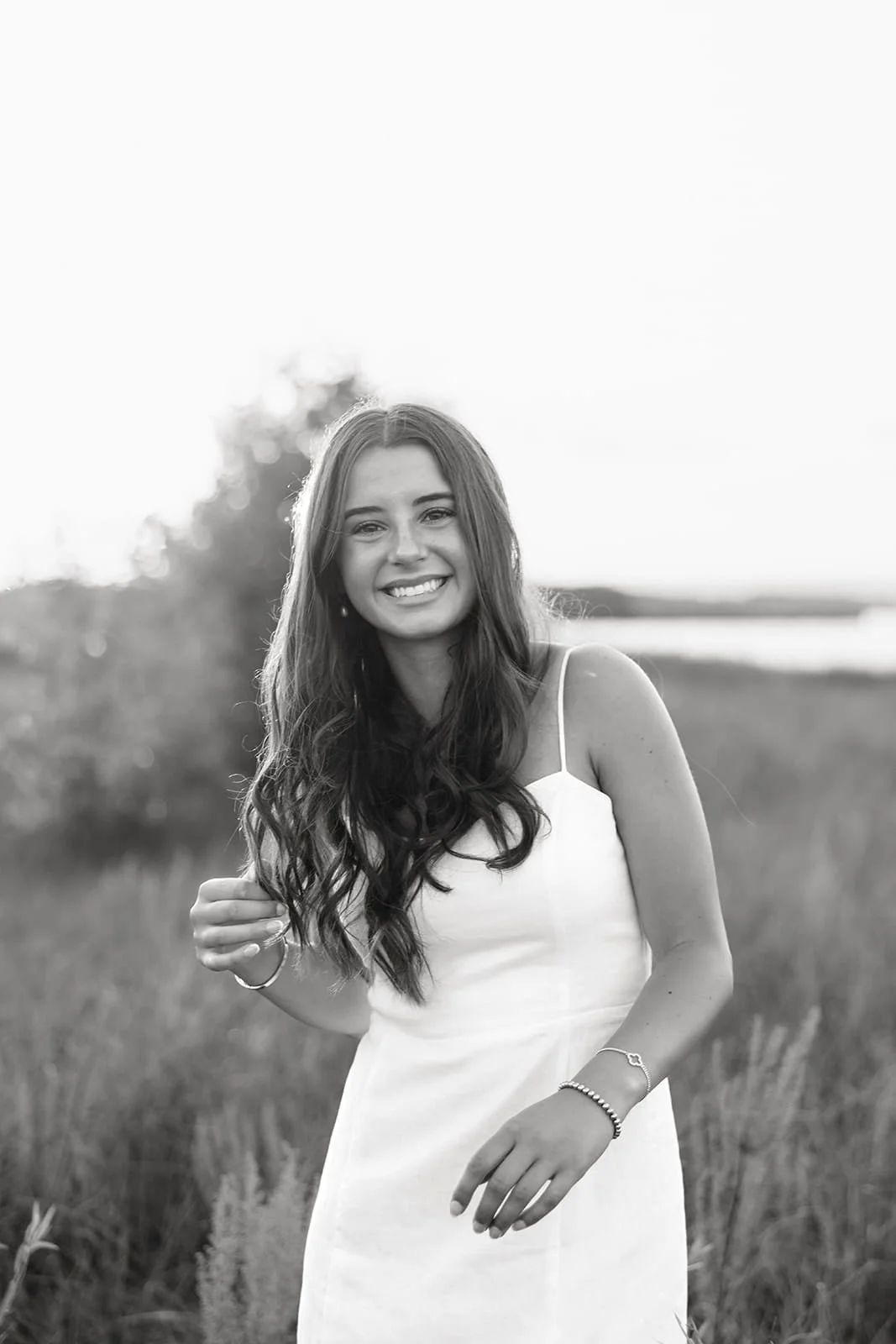 A young woman with long wavy hair smiling, standing outdoors in a field, wearing a sleeveless white dress and jewelry, in black and white.