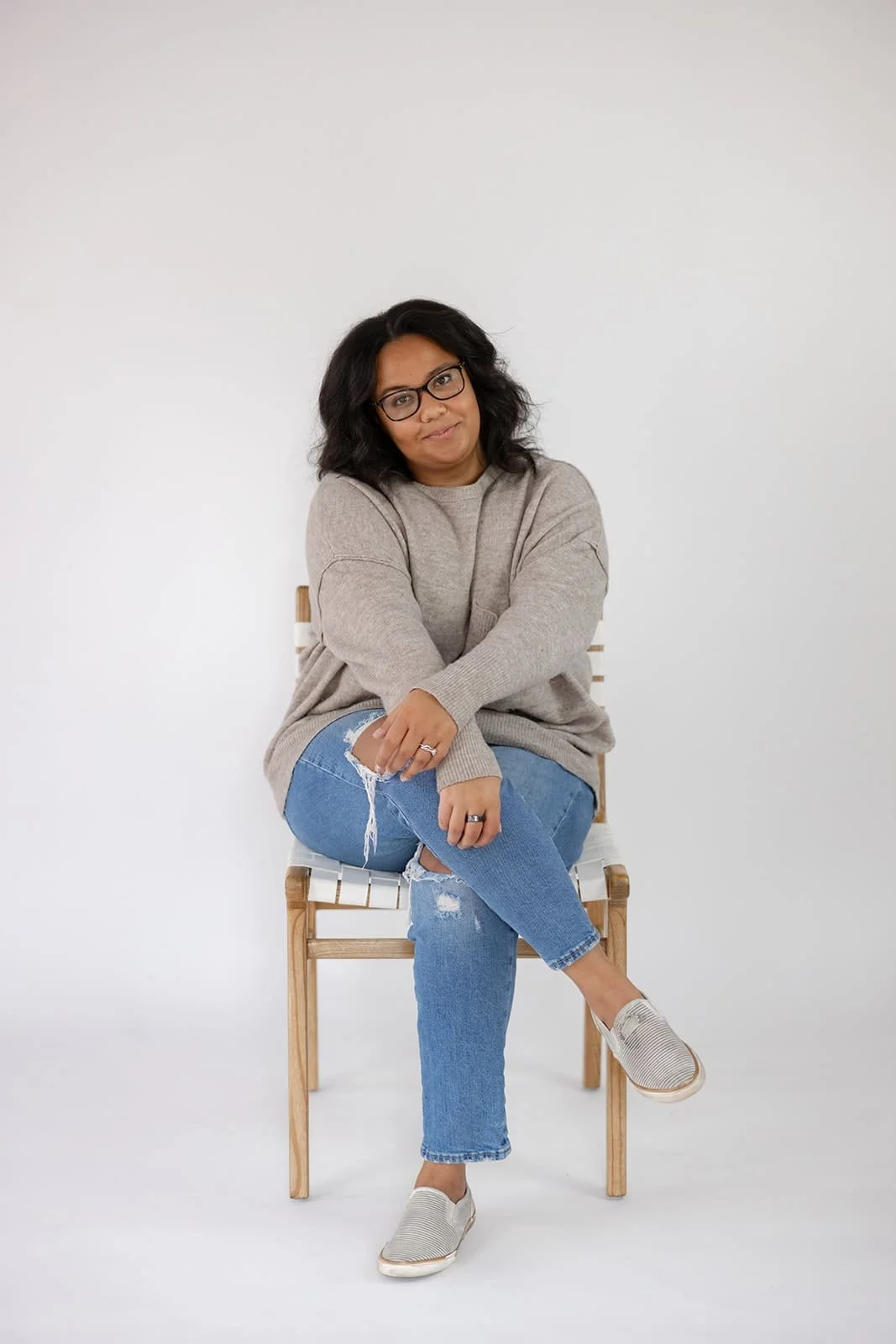 Woman with glasses sitting on a wooden chair against a plain white background, wearing a beige sweater, ripped jeans, and slip-on shoes.