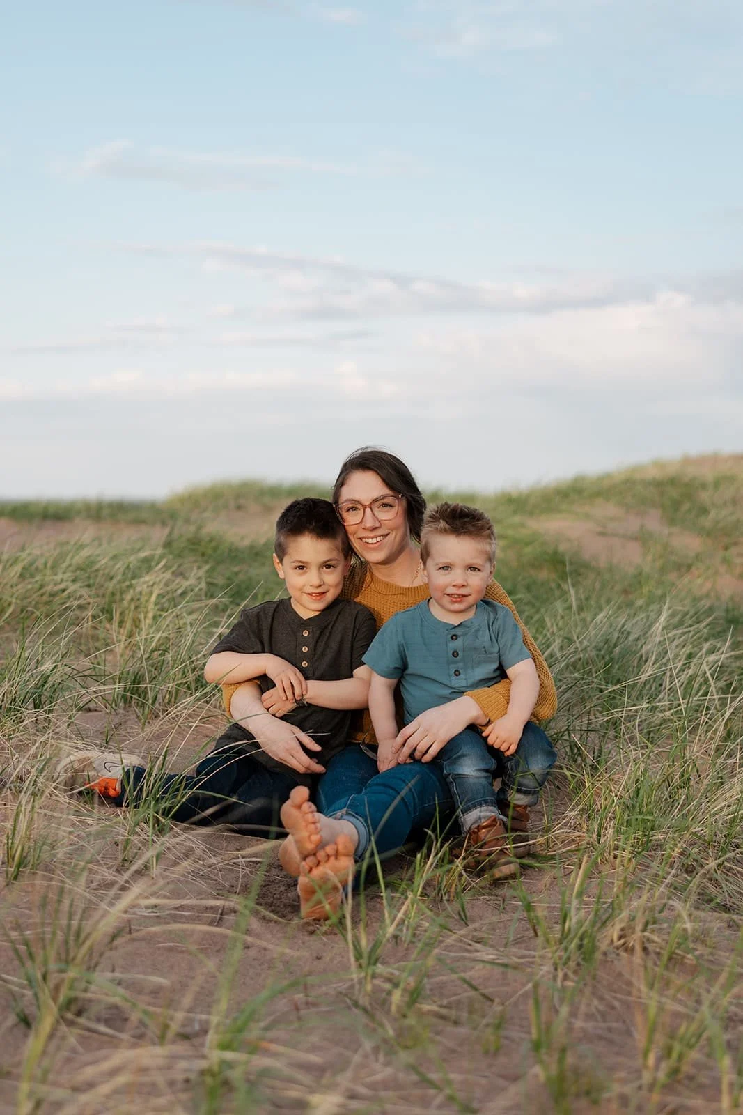 A woman sitting on the sand with two young boys, in a grassy outdoor setting under a partly cloudy sky.
