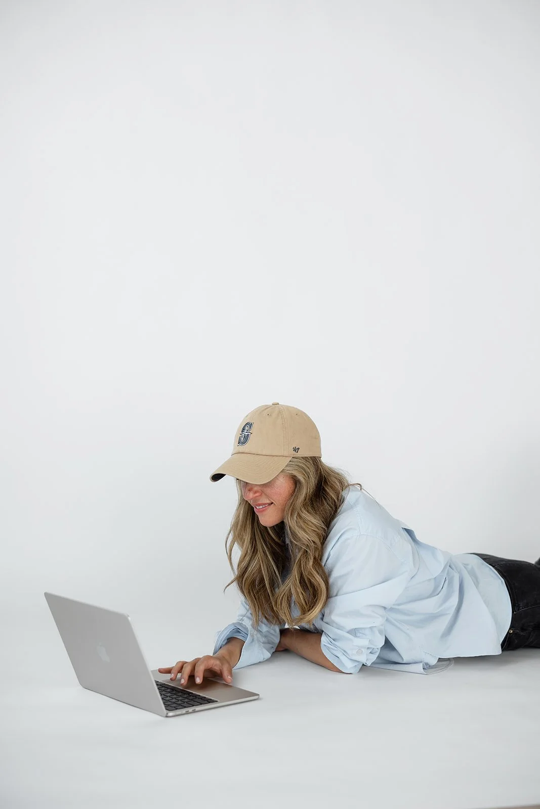 Woman lying on the floor, wearing a beige baseball cap and light blue shirt, working on a silver laptop against a plain white background.