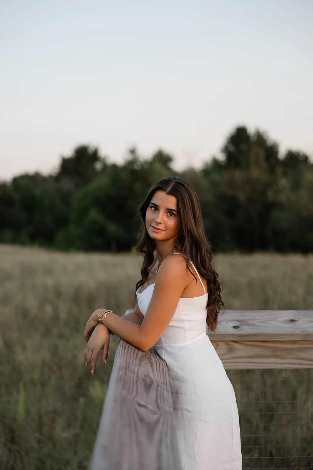 A young woman with long brown hair wearing a white dress, leaning on a wooden fence in a grassy field during sunset.