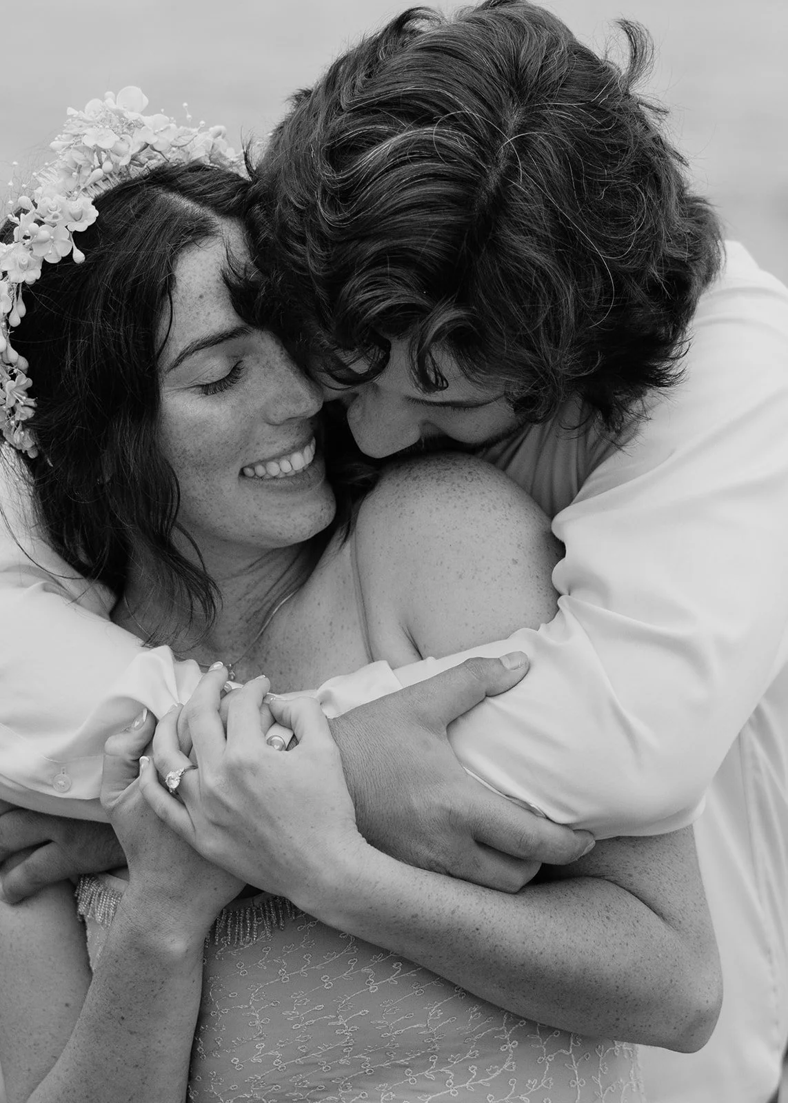 A woman with dark curly hair and a floral headband is smiling while hugging a man with wavy hair. They are embracing tightly with their faces close together, and the woman is showing her wedding ring.