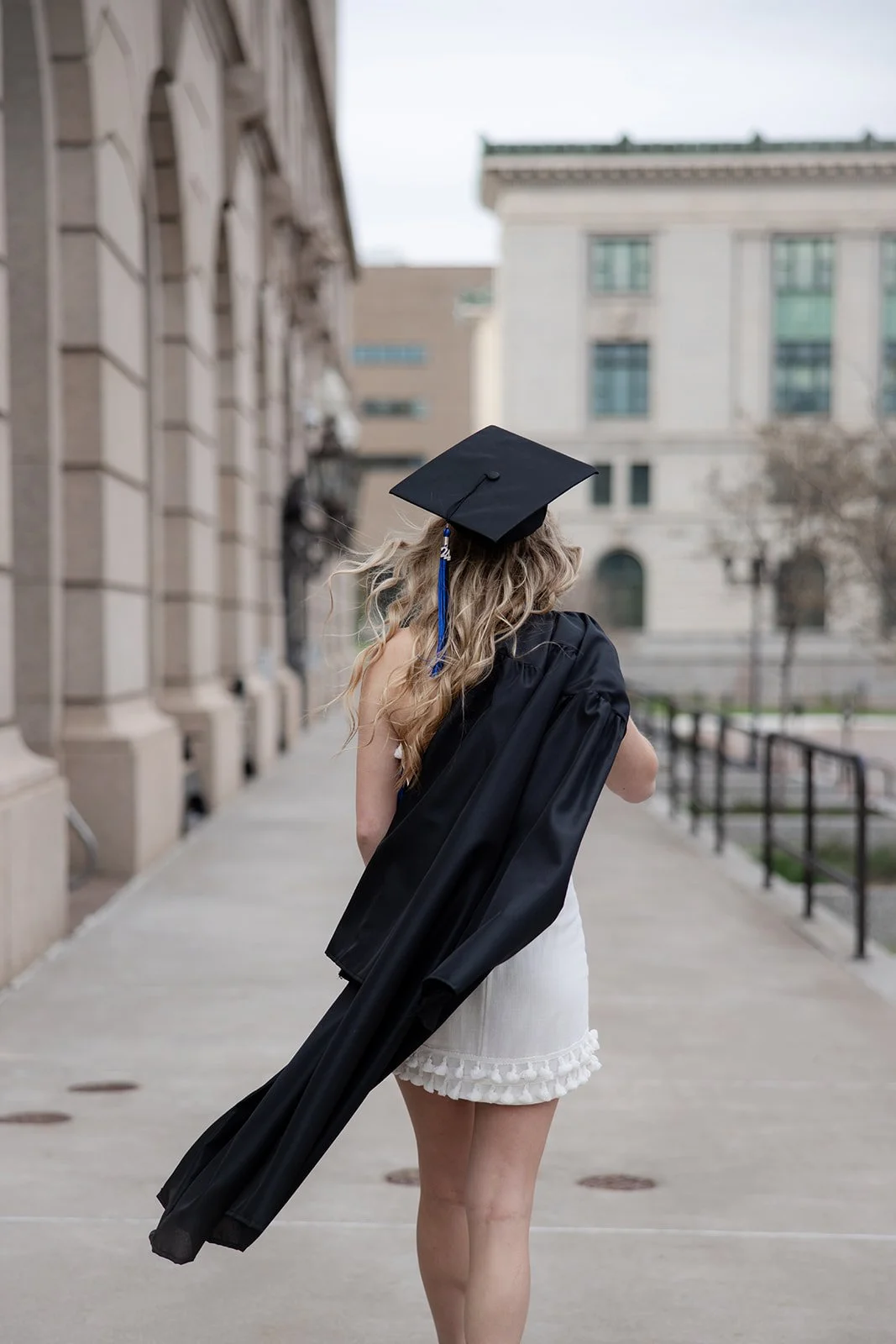 A woman in a graduation cap and gown holding a stole on her shoulder, walking along a city sidewalk with classical buildings in the background.