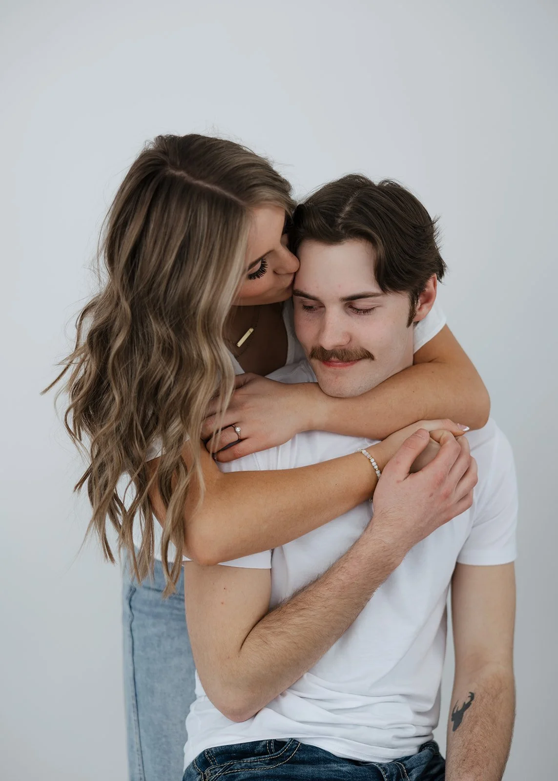 A woman with long wavy blonde hair kissing a man with dark hair and a mustache on the head, as they embrace each other affectionately.