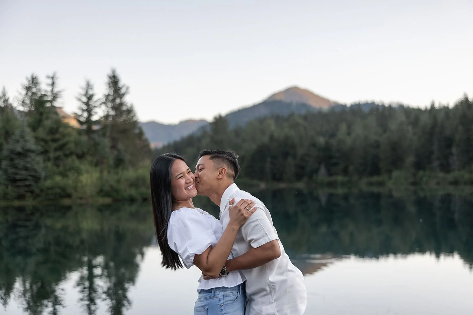 A couple hugging and kissing near a body of water with trees and mountains in the background during sunset.