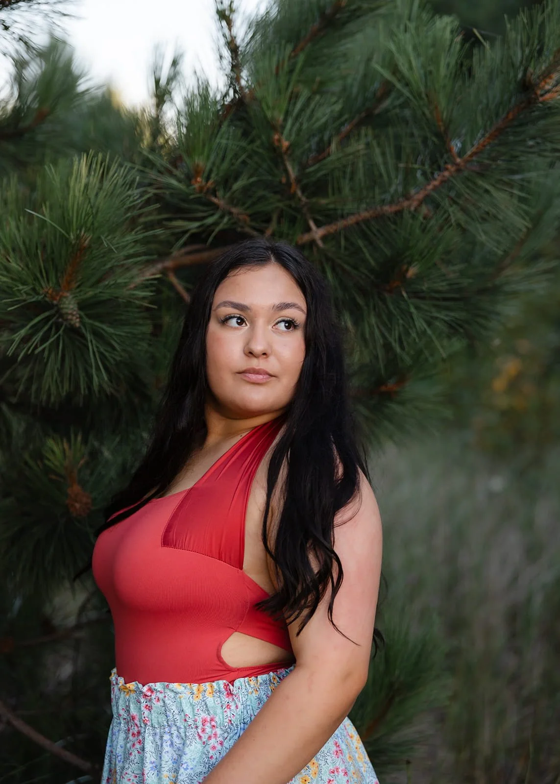A woman with long black hair and tan skin standing outdoors in front of a green pine tree, wearing a red sleeveless top and a floral skirt.