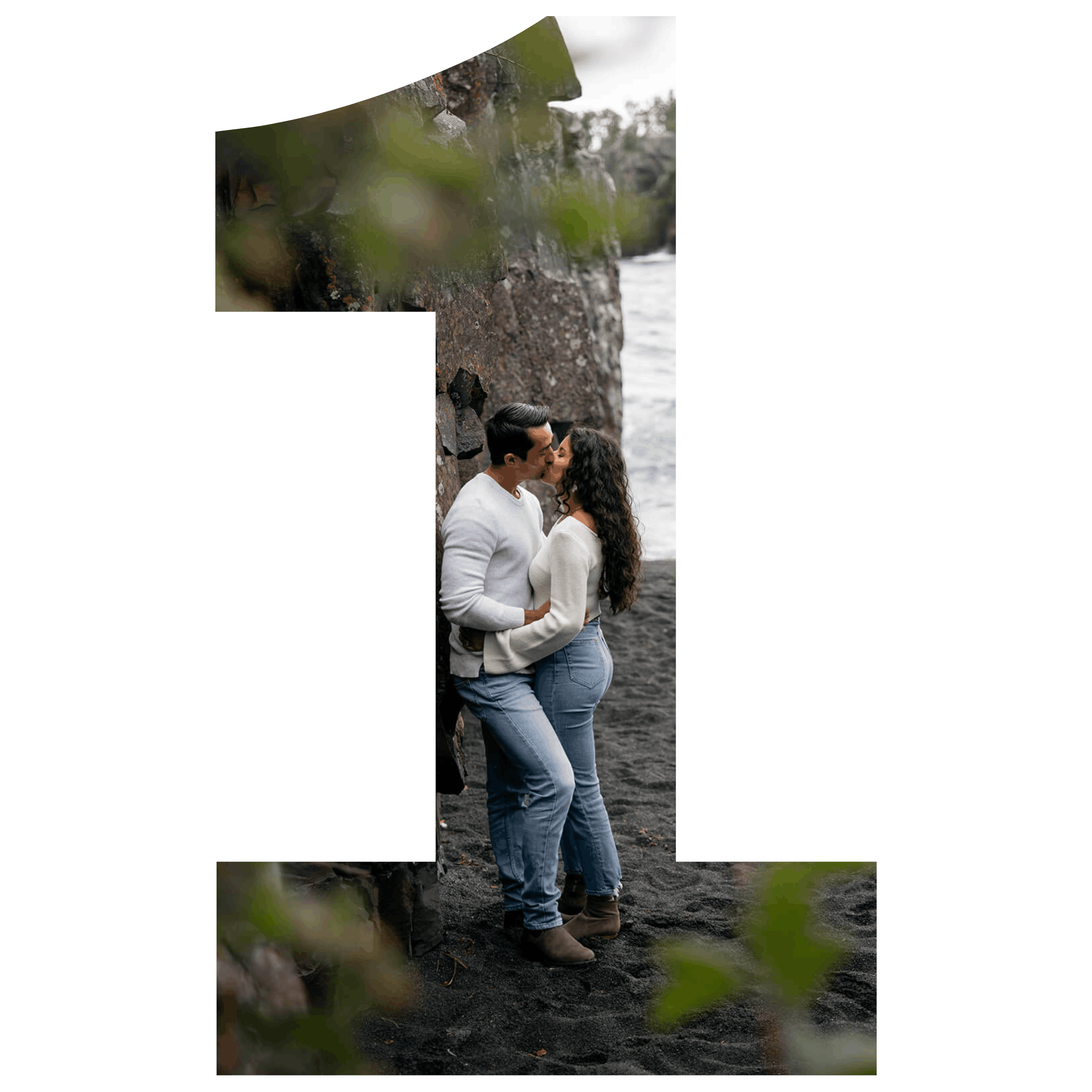 A couple sharing a kiss on a black sand beach near a rocky cliff, with green leaves in the foreground.