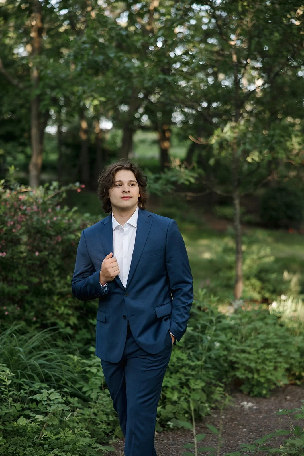A young man in a blue suit and white shirt walking outdoors in a green park or garden with trees and bushes, looking off into the distance.