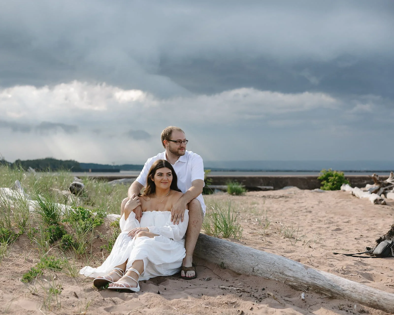 A couple sitting on a sandy beach beneath stormy skies, with green plants and driftwood around them, and water in the background.