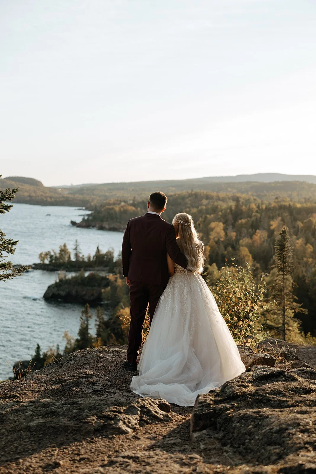 Bride and groom standing on a rocky hill overlooking a river and forest, with a sunset sky in the background.