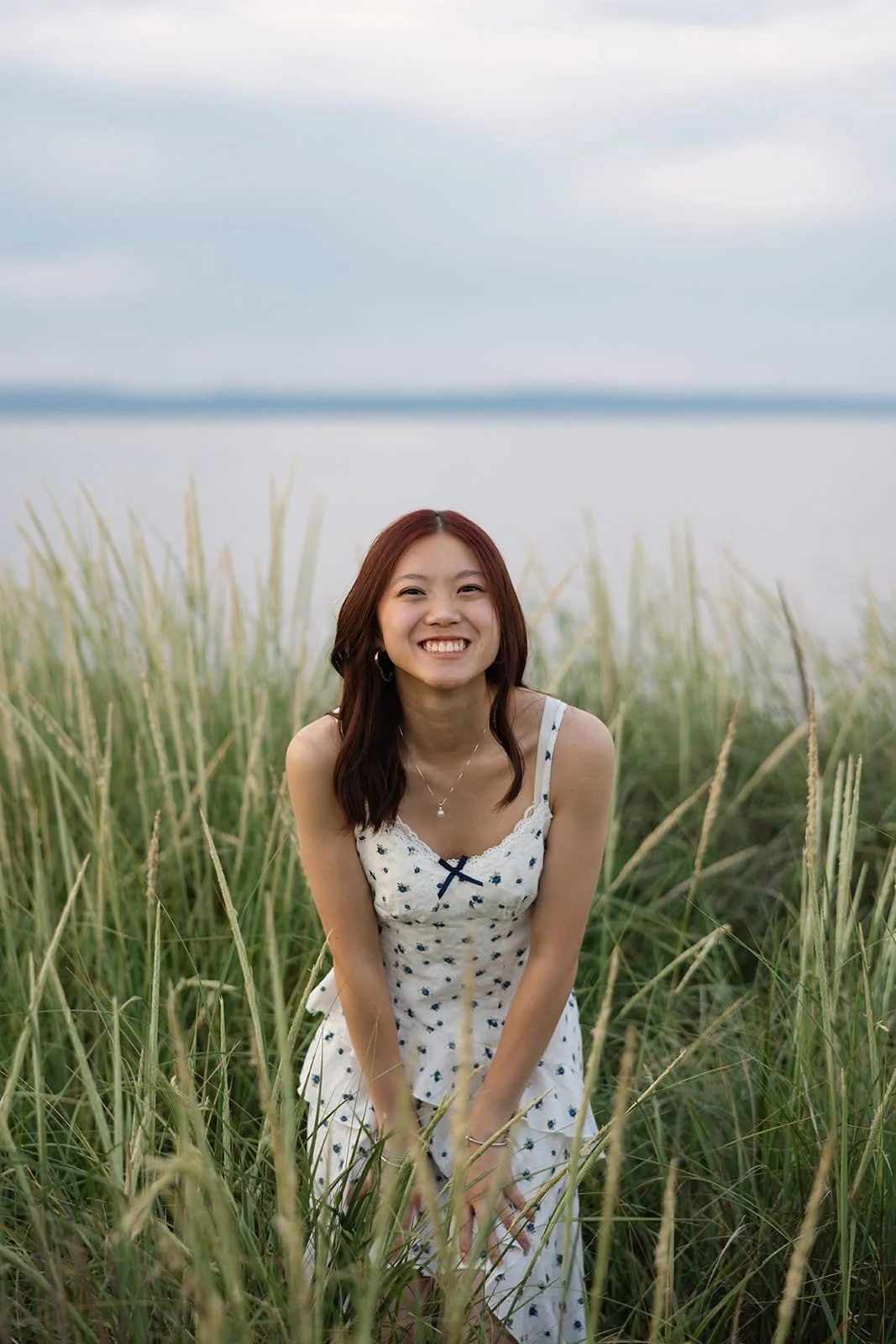 A young woman with shoulder-length brown hair, wearing a white dress with black polka dots and a small black bow, smiling while standing among tall green grass near a body of water with a cloudy sky overhead.