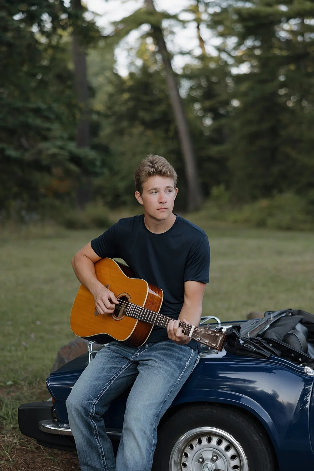 A young man sitting on the hood of a blue vintage car, playing an acoustic guitar outdoors in a wooded area.