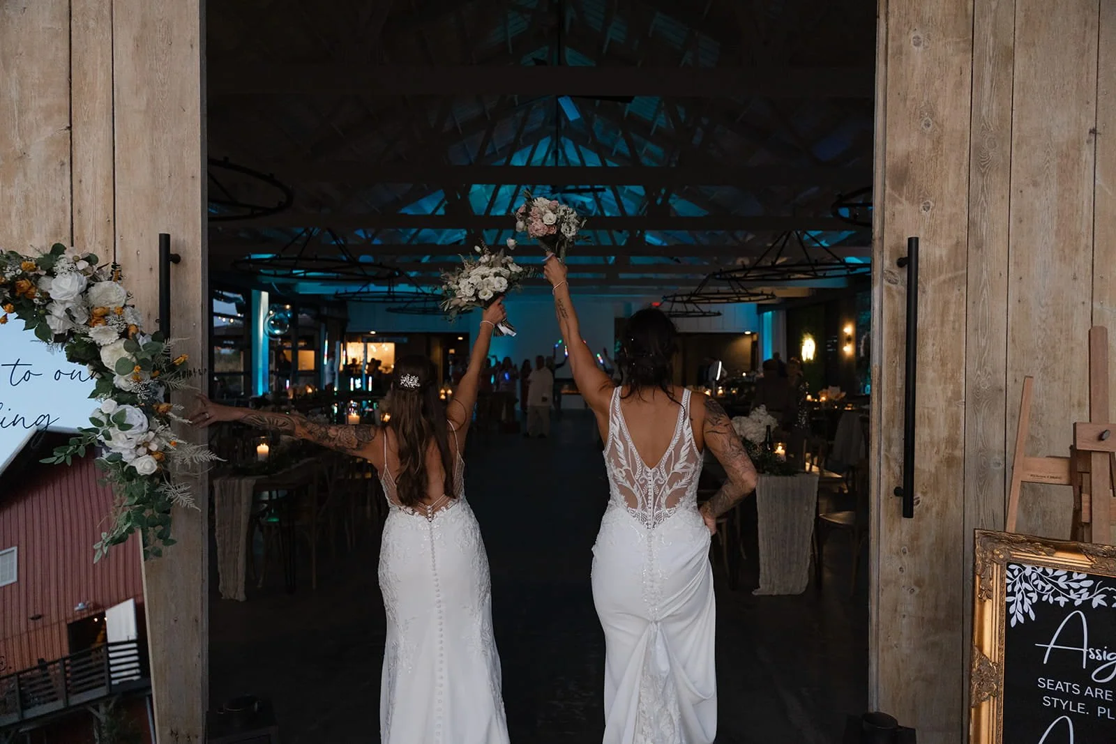 Two brides in wedding dresses hold bouquets and walk into a wedding reception hall, raising their bouquets in celebration.