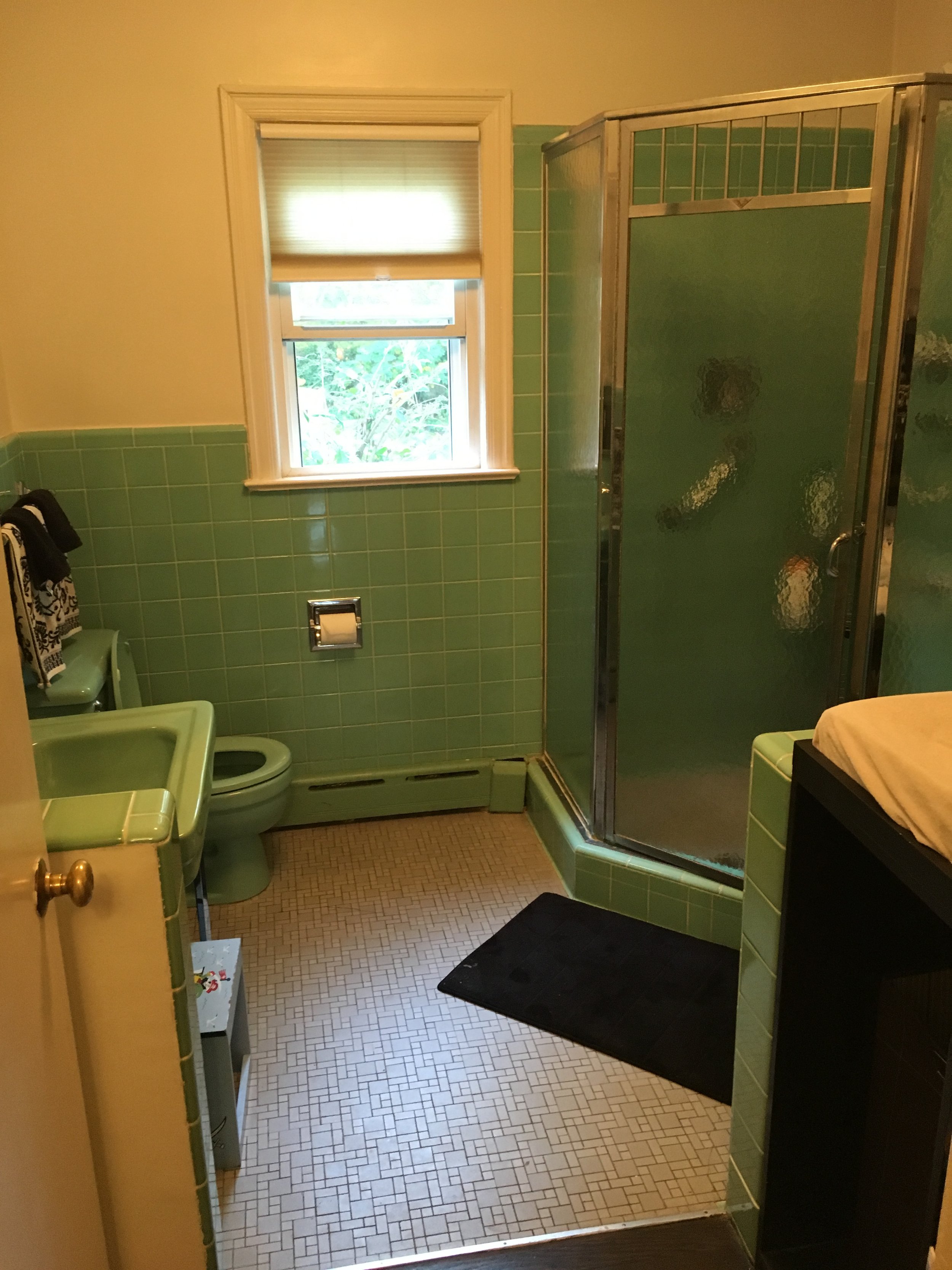 A vintage bathroom with green tiles, a green toilet, a green sink, a black rug, a window with a beige shade, and a green-tiled shower stall with frosted glass.