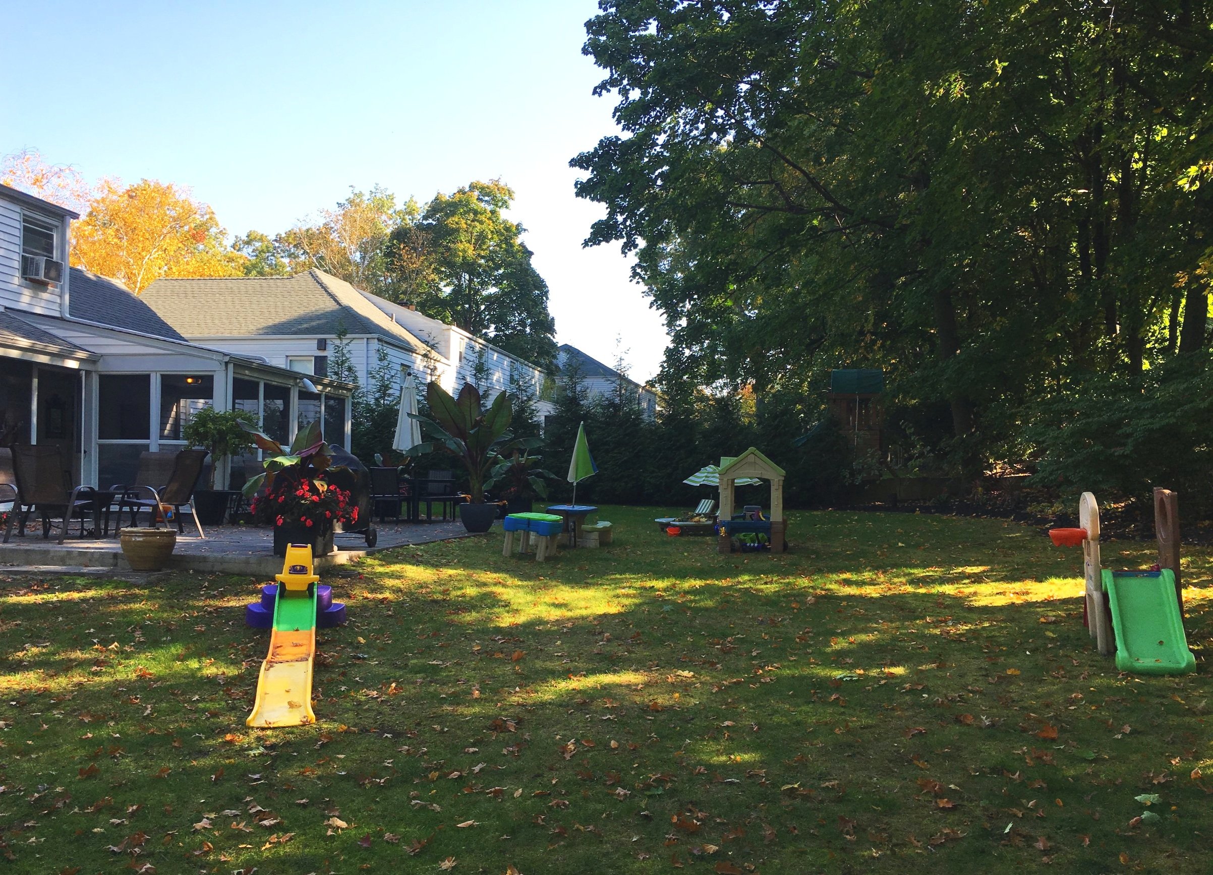 Residential backyard with grass, trees, children’s slides, outdoor furniture, and a house with a porch.