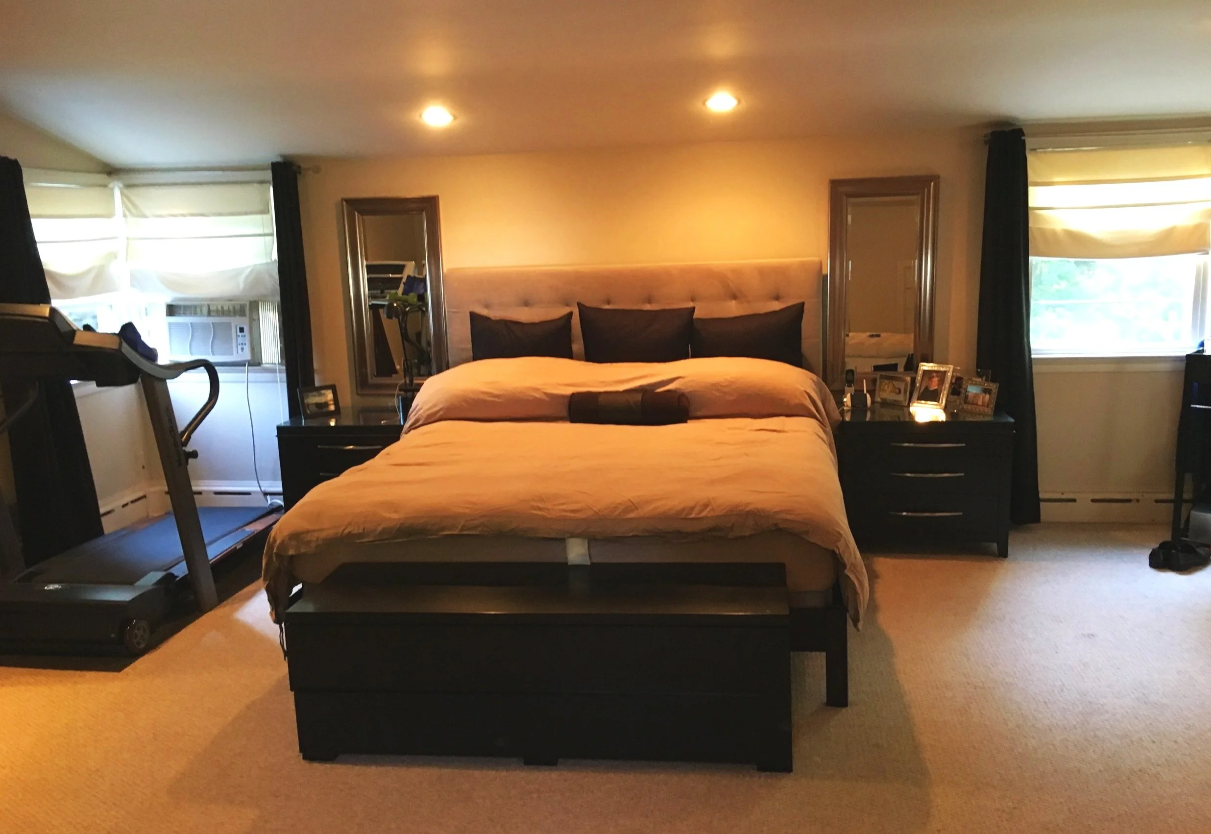 A bedroom with a large bed, black pillows, dark furniture, windows with curtains, a treadmill, and personal photos on nightstands.
