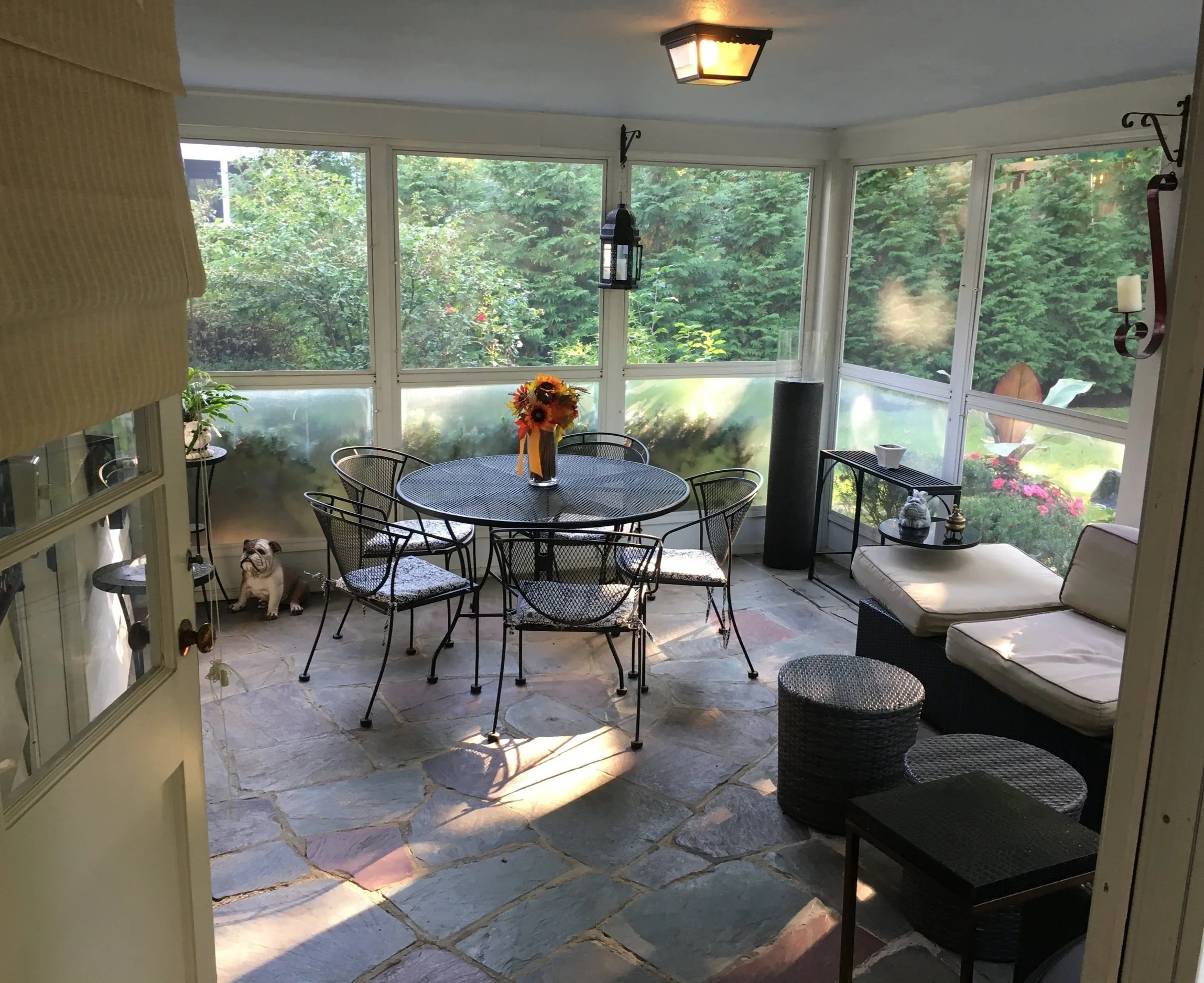 Sunroom furnished with a round metal dining table with a sunflower centerpiece, six chairs, a wicker sofa with cushions, a small black side table, and decorative items. Large windows show lush green garden outside, and the floor is made of stone tiles.