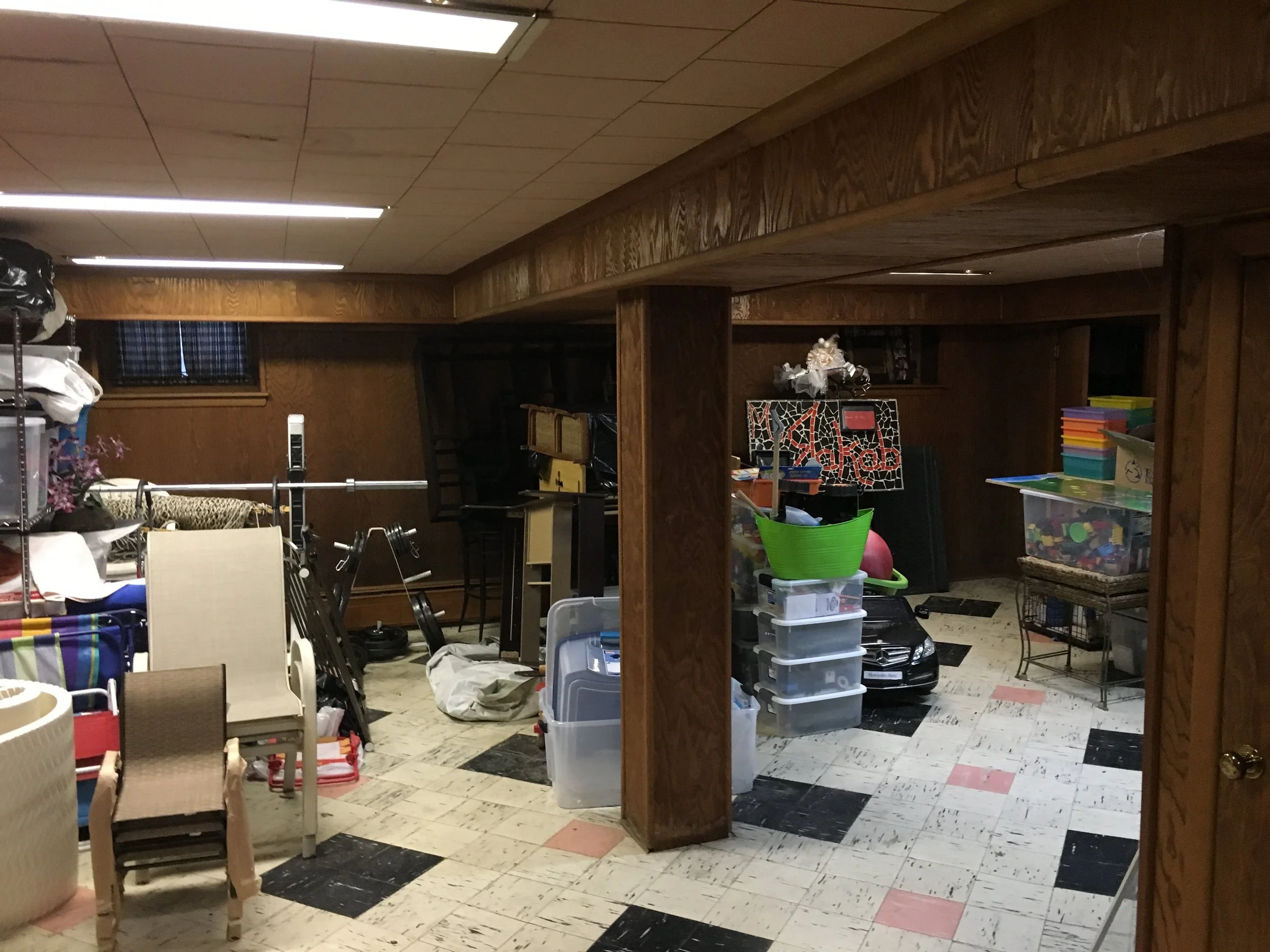 A cluttered basement room filled with various household items including plastic storage bins, furniture, toys, and miscellaneous objects on the floor and shelves. The room has wood-paneled walls, a drop ceiling, and a black-and-white tiled floor.