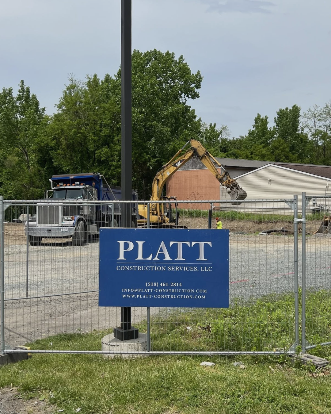 Construction site with a yellow excavator and a large blue dump truck behind a metal fence. A construction worker is visible in the background, and a blue sign for Platt Construction Services, LLC is on the fence.