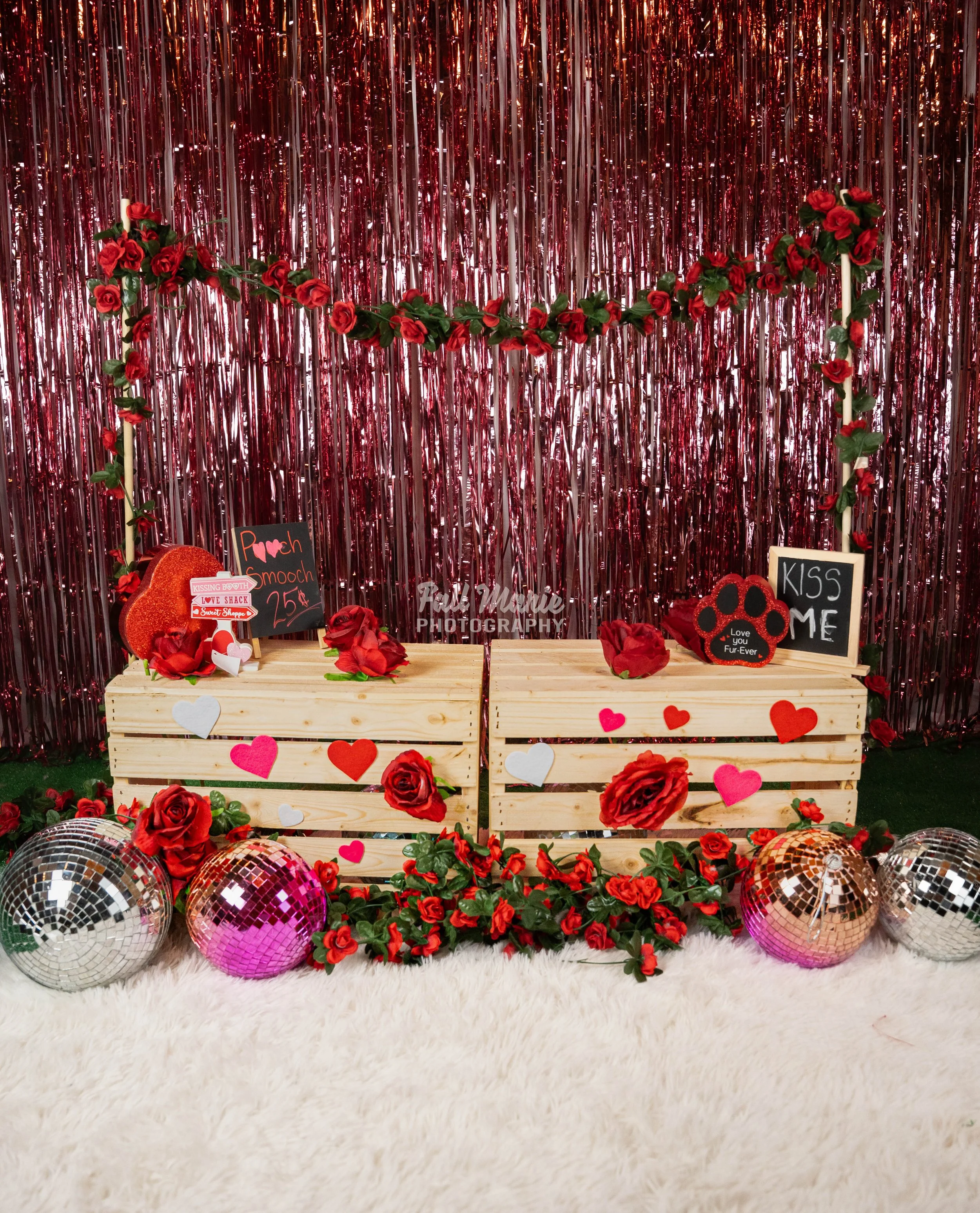 Valentine's Day-themed display with two wooden crates decorated with red and pink paper hearts and red roses, surrounded by artificial flowers and three disco balls, set against a pink metallic fringe backdrop with a floral garland.