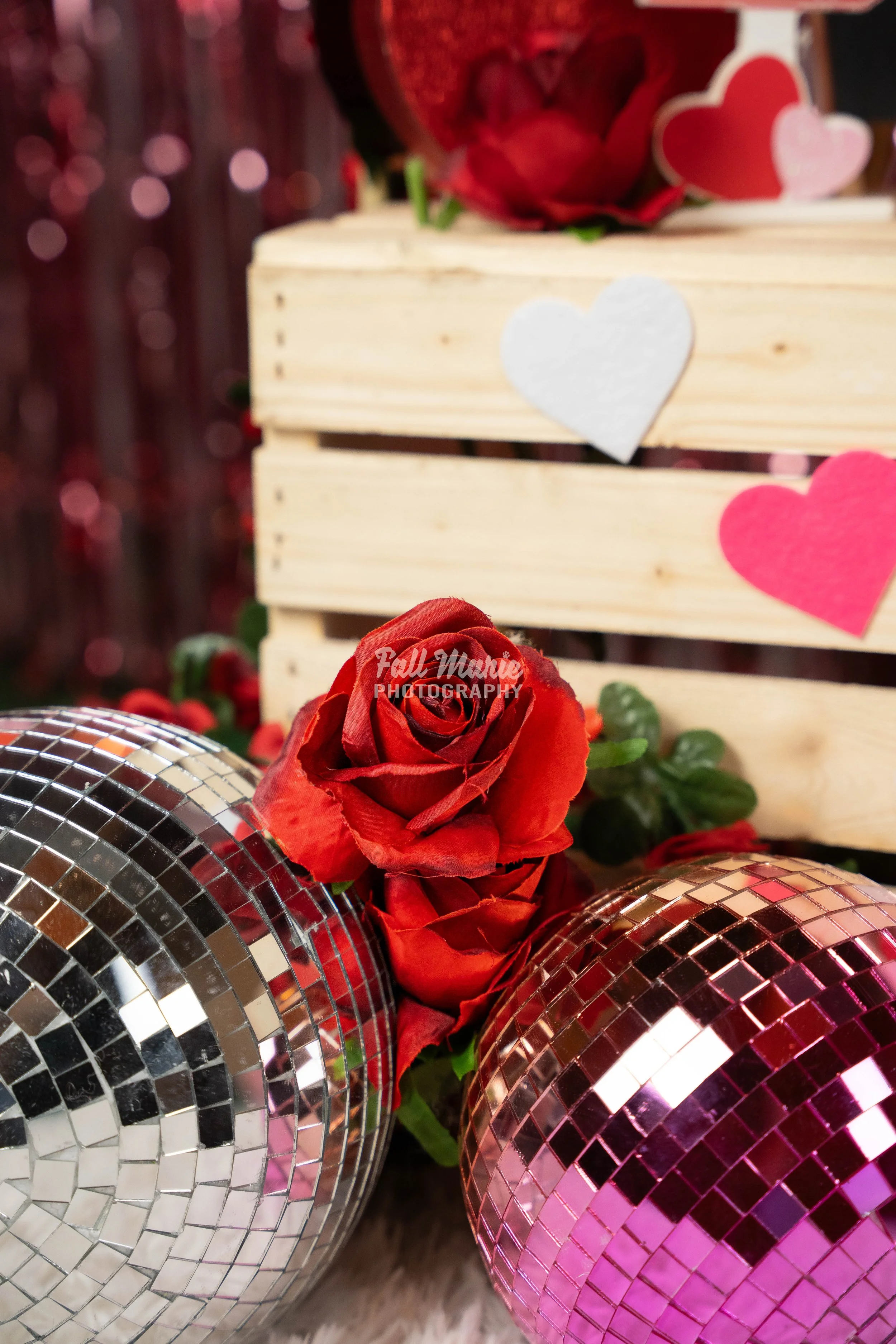Red roses, two disco balls, and multicolored paper hearts on a wooden crate, celebrating love and Valentine's Day.