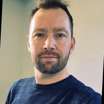 A man with short dark hair and a beard takes a selfie, wearing a dark t-shirt against a plain background.