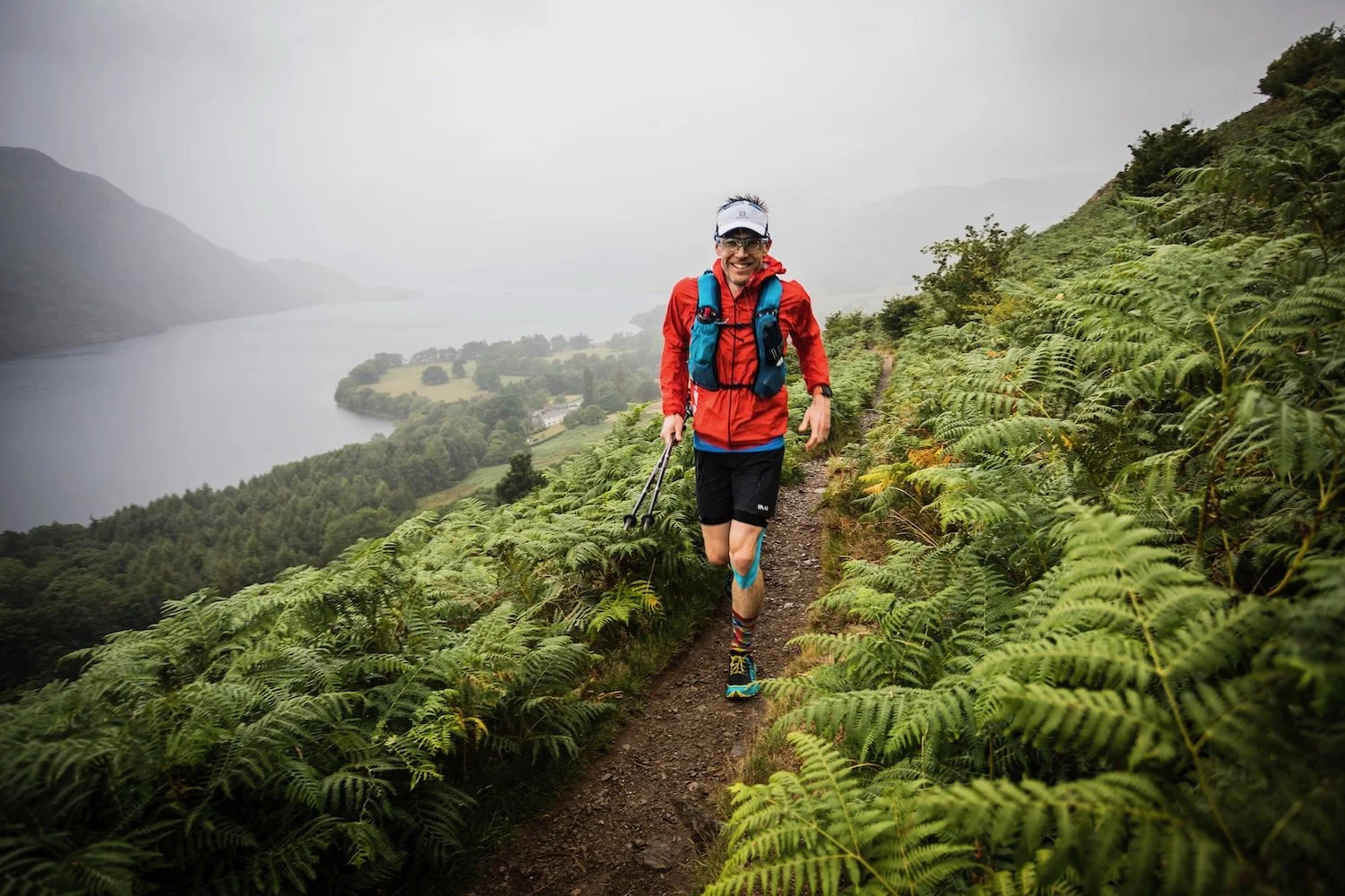 Man running on a trail surrounded by green ferns, with a lake and mountains in the misty background.