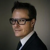 A man with short dark hair, wearing black-framed glasses, a dark suit, and a white shirt, posing for a professional portrait against a dark background.