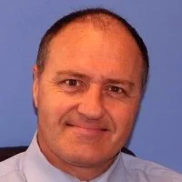 Portrait of a middle-aged man with short dark hair, wearing a light blue shirt, smiling, against a blue background.