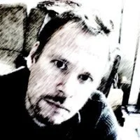 Black and white photo of a man with short hair, a beard, and glasses, looking at the camera.